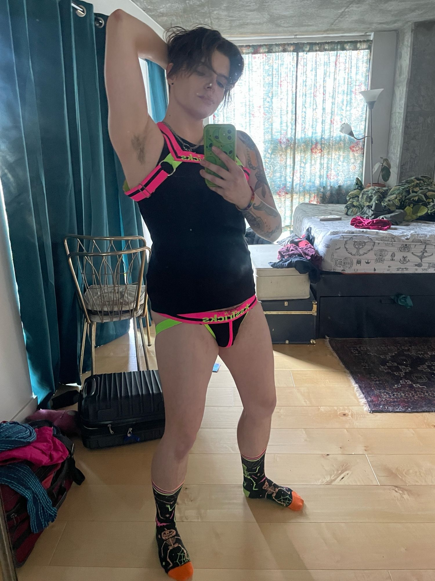 Micki wearing a black tank top with a neon pink and green harness and matching jock strap. Also some neon skeleton socks