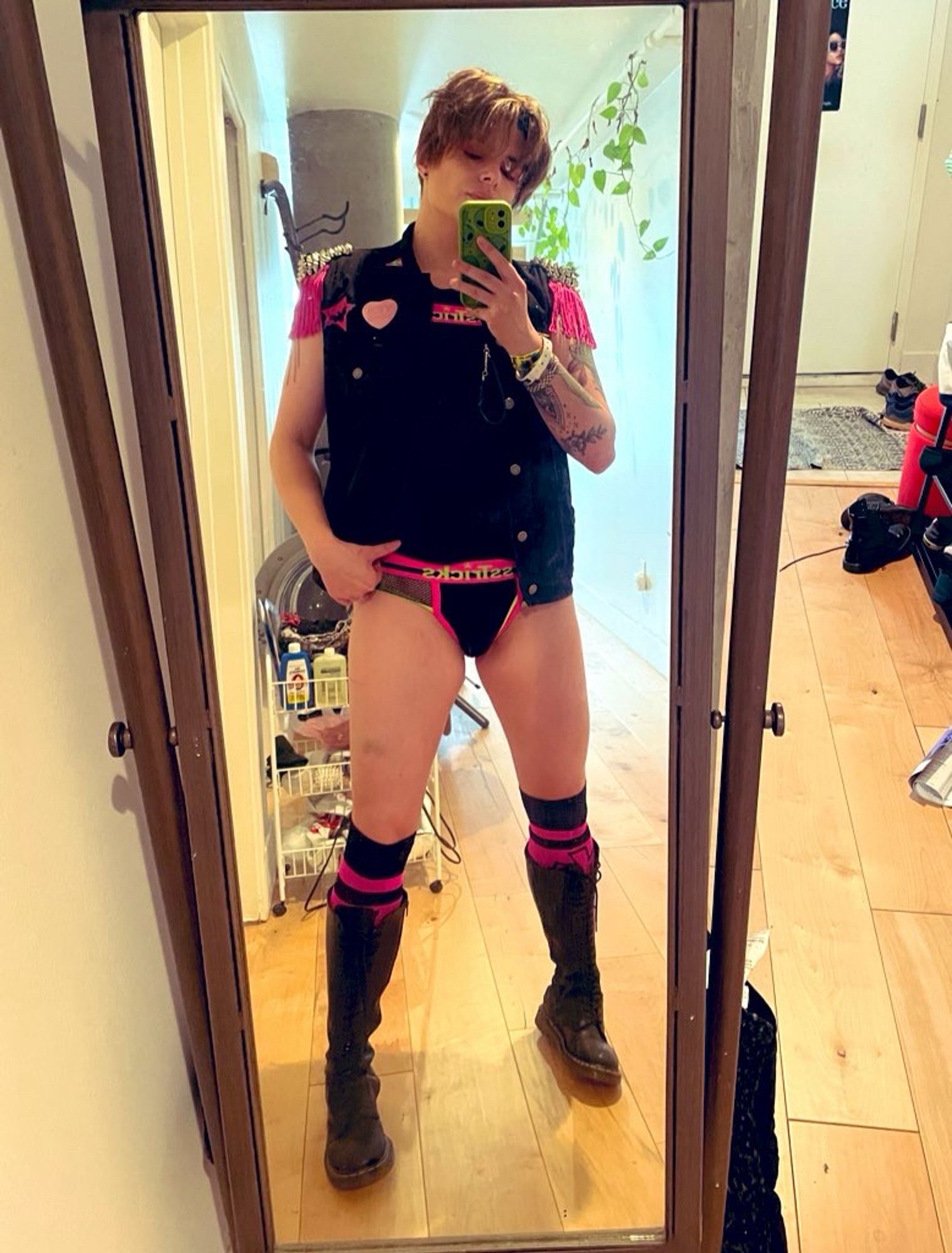 Micki wearing all black with hot pink accents. Knee high boots with fishnet briefs, a punk vest with hot pink fringe and buttons saying queer and yes homo