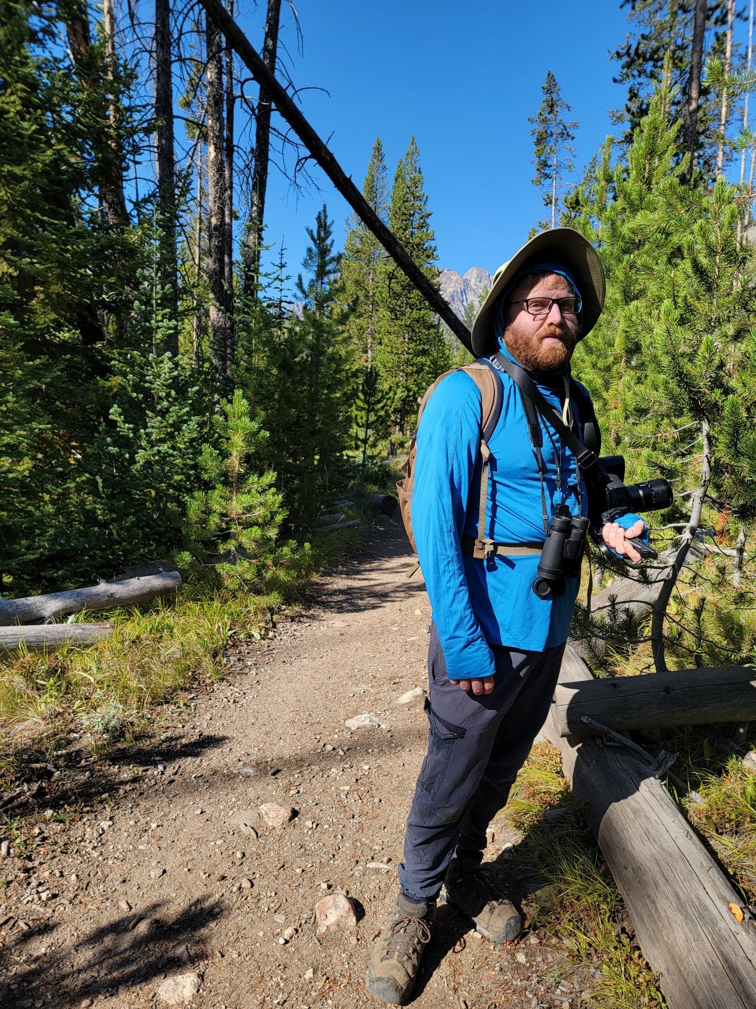 I'm standing in a forest, on a trail. I have a big hat on my head and heavy black binoculars on my chest, and large camera in my arm.