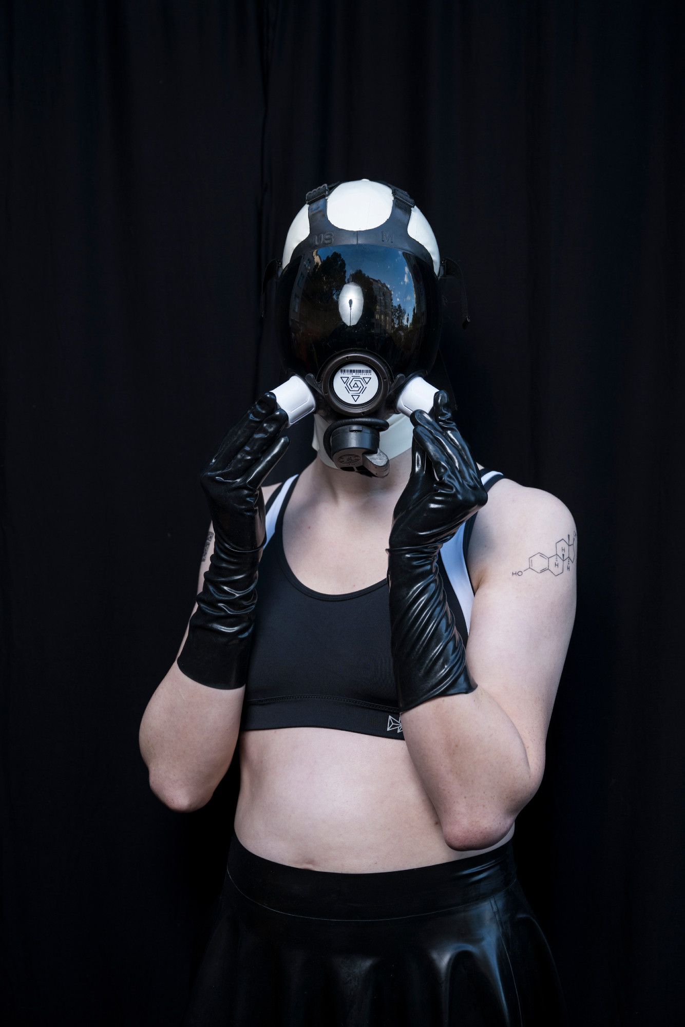 Dana in its drone outfit, latex clad hands touching the inlets of its gasmask