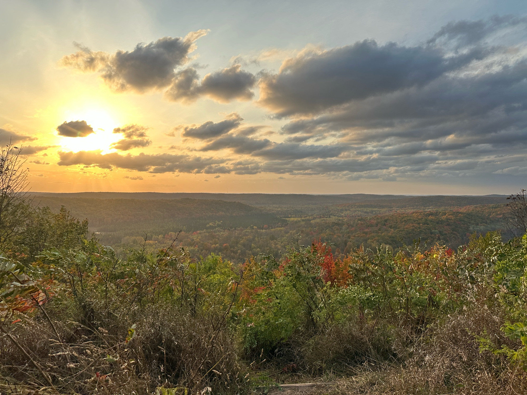 A picture from a tall hill, looking out over a valley. The trees are turning color, so the tree cover is a mix of green, yellow, orange and red. The sun is in the top left, about an hour from setting, the sky a bright yellow and orange