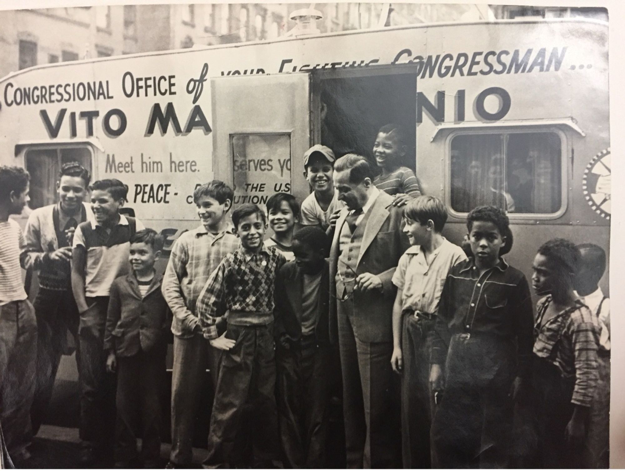 Rep. Marcantonio at his office, a trailer, with neighborhood children, ranging in age, from very young to teenagers. They are standing, some posing, some talking, with Marcantonio. The door to the trailer is open with some children standing inside the door frame, laughing. Marcantonio looks down at the child to his right, smiling warmly, with their arms around each other. The trailer reads "Congressional Office of your...Congressman," "Vito Ma....nio, "Meet him here," "PEACE," "Serves you..." Ellipsis in text signals what is blocked by the open door. Buildings line the top frame of the photo, behind Marcantonio's office.