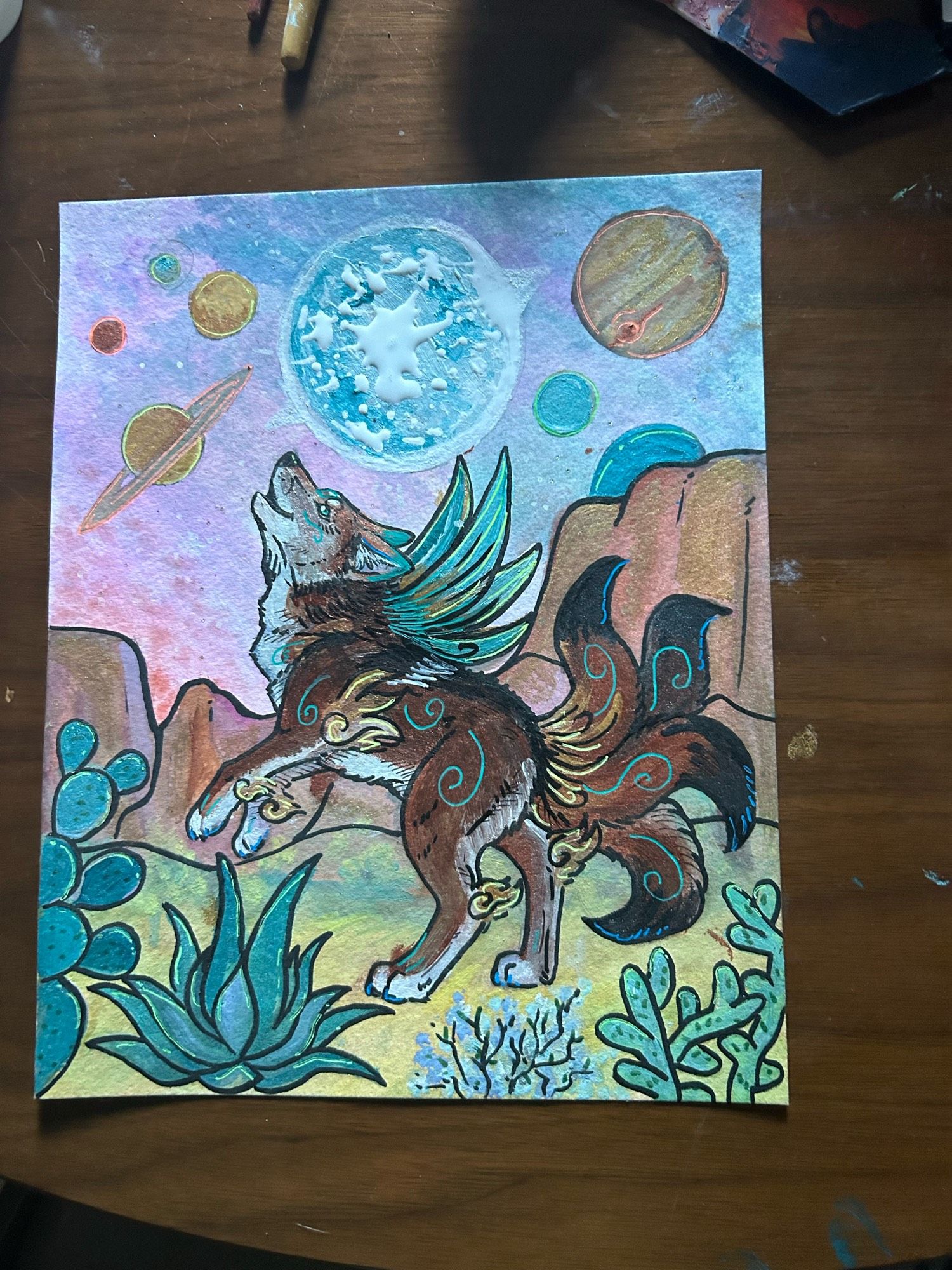 Watercolor and ink painting of a coyote standing on its hind legs and looking up. It has four tails, blue wings, and blue swirls on its fur. The background is a desert, with the sky full of planets and stars