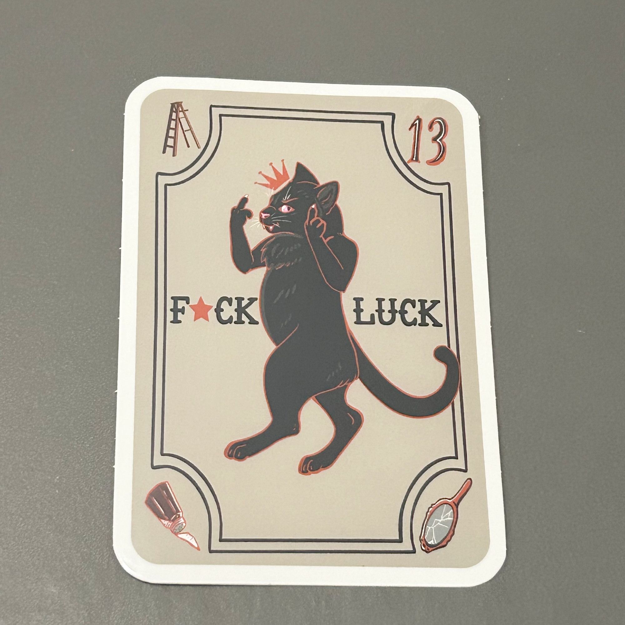 Sticker of a black cat holding up its middle "fingers" and walking on two legs. It says "Fuck Luck". It is in the shape of a playing card. In the corners, there is a ladder, the number 13, spilled salt, and a broken mirror.