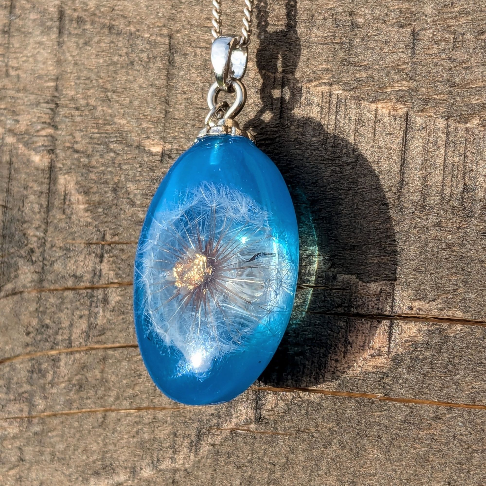 Pendentif oval en résine bleue avec un mini pissenlit au centre, dont le cœur est en feuille d'or. Une breloque en feuille le complète.