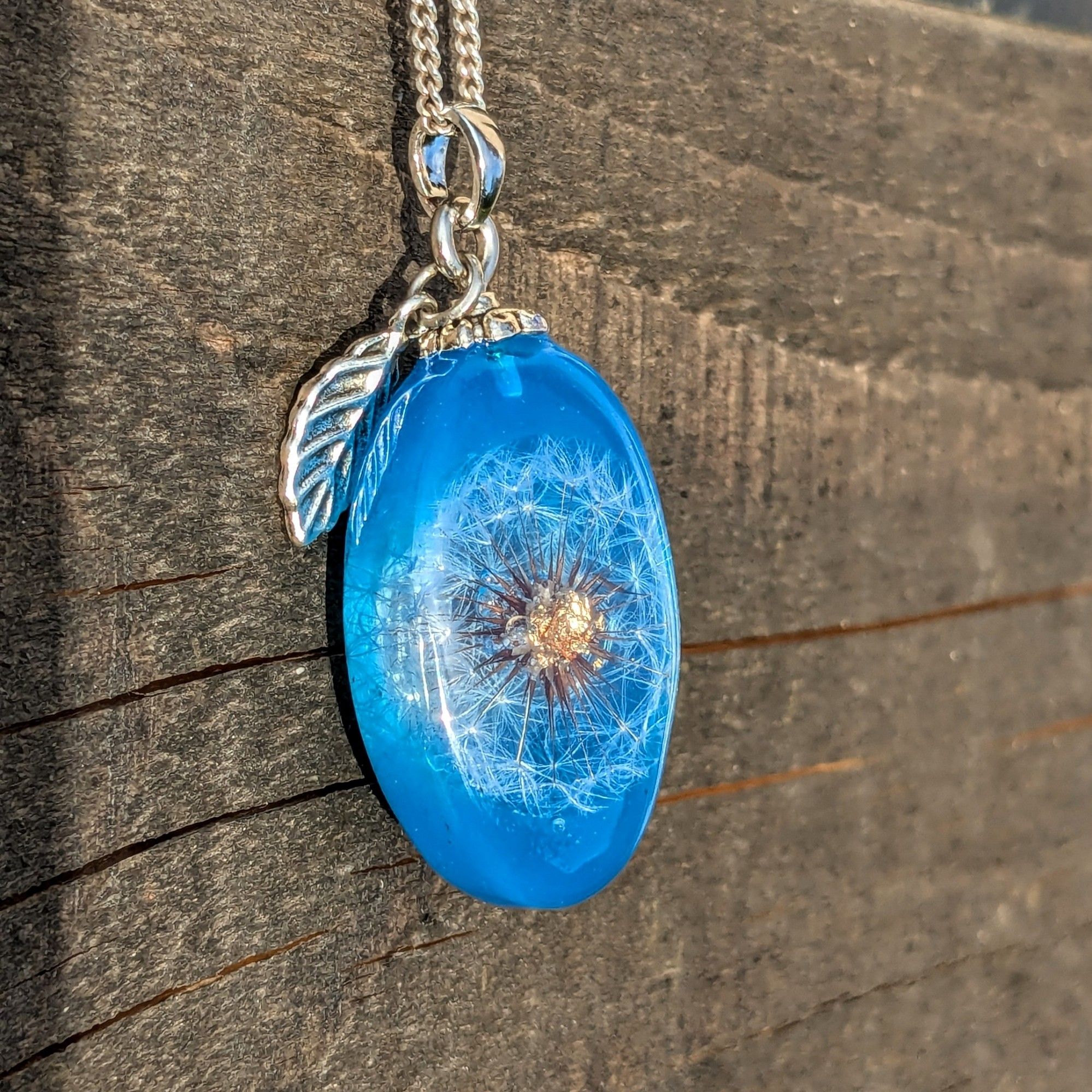 Pendentif oval en résine bleue avec un mini pissenlit au centre, dont le cœur est en feuille d'or. Une breloque en feuille le complète.