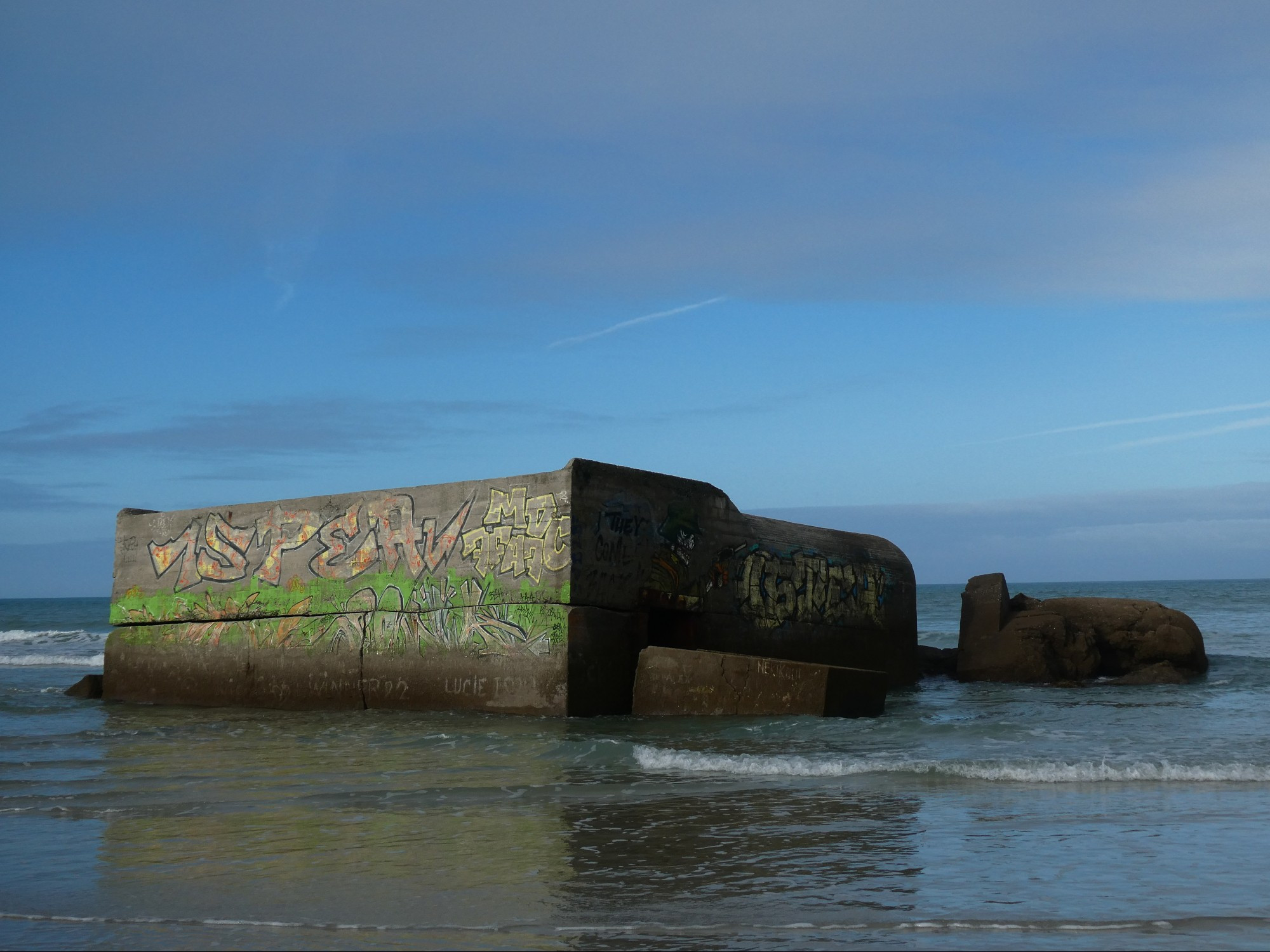 Graffitied remains of concrete sea defences. High tide lapping around it.