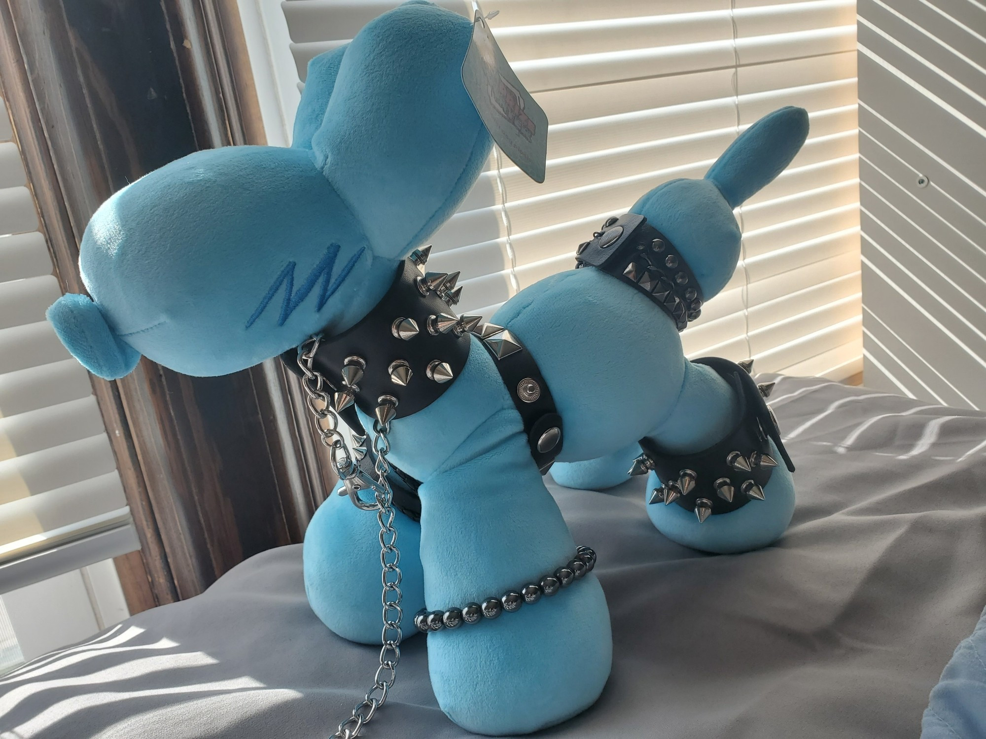 A picture of a plush blue balloon animal, wearing tons of goth spiky armbands and with a chain collar. The plush is made by https://theprizecounter.com/