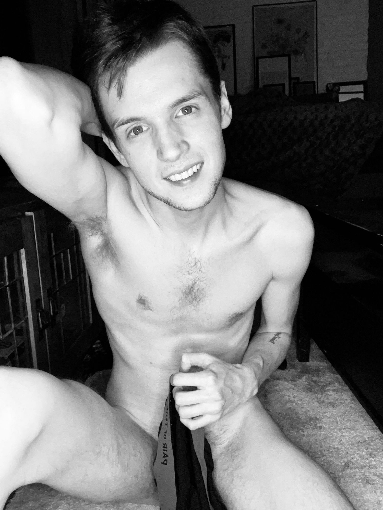 A black and white photo of me with my legs spread holding some underwear in front of my cock to provide a tease