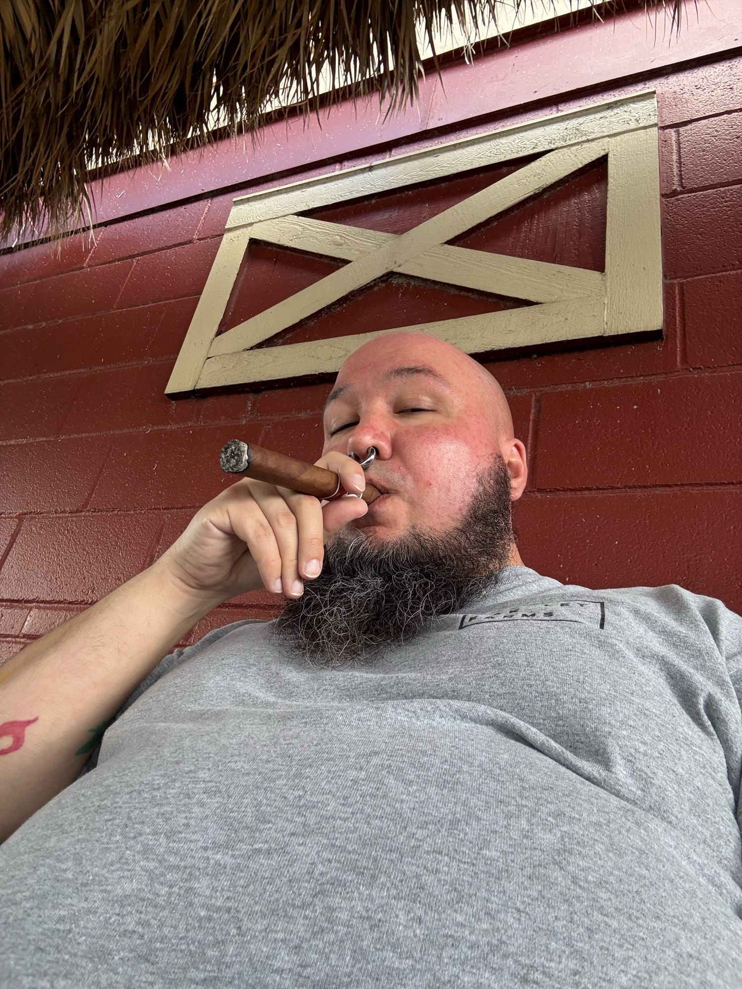 Selfie of a bald man with a black and grey beard wearing a grey shirt and metal septum ring. Puffing on a cigar.