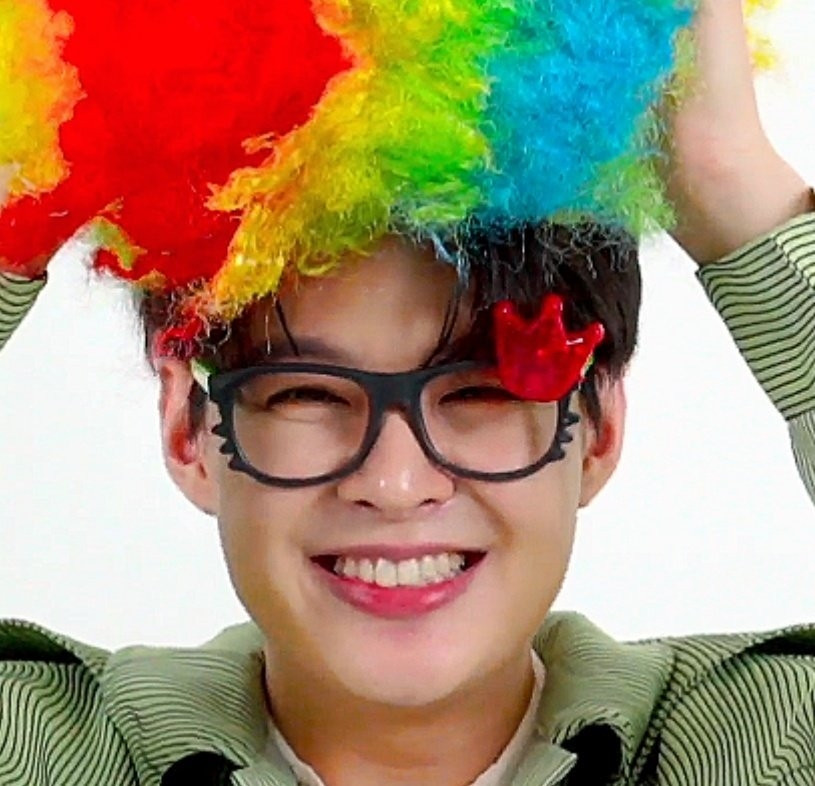 Saint holding a colorful clown wig on his head with both hands. He's wearing fake glasses with large black frames and a little red crown pin on them. His smile is dazzling, eyes narrow with joy.