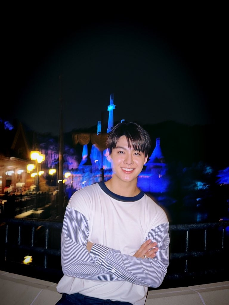 Saint with a joyous smile and his arms crossed in a relaxed way. It's night and there's a blue lit castle in the far background and some yellow lights from lightposts on the left 