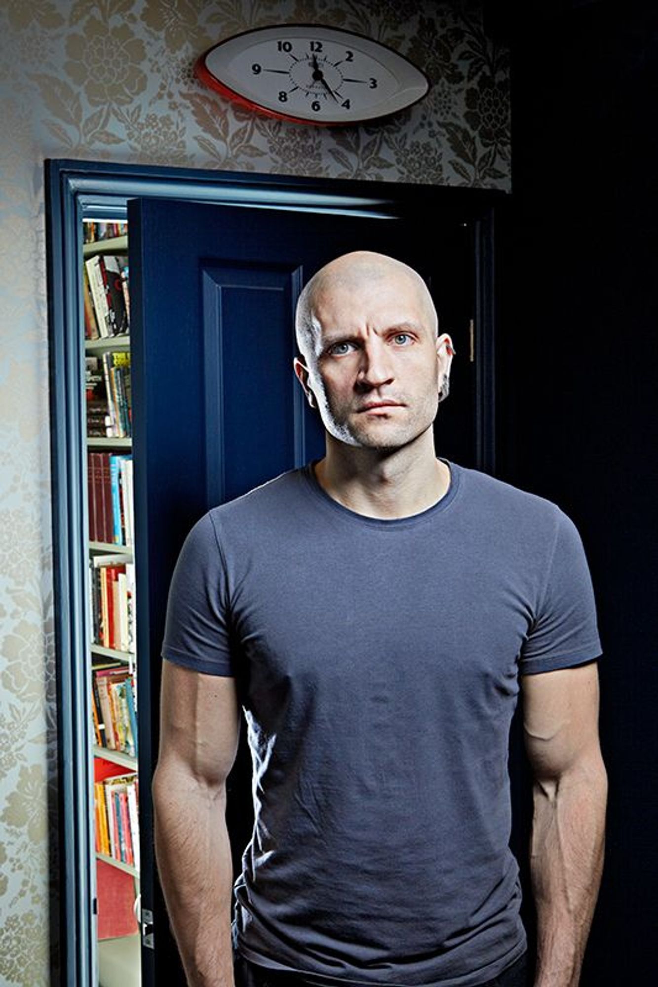 photograph of a shaved-head man with many earings standing in front of a door into a room full of books. he's wearing a grey t-shirt, and is pretty jacked. big ol poppin' bicep vein going on. it's china mievile!