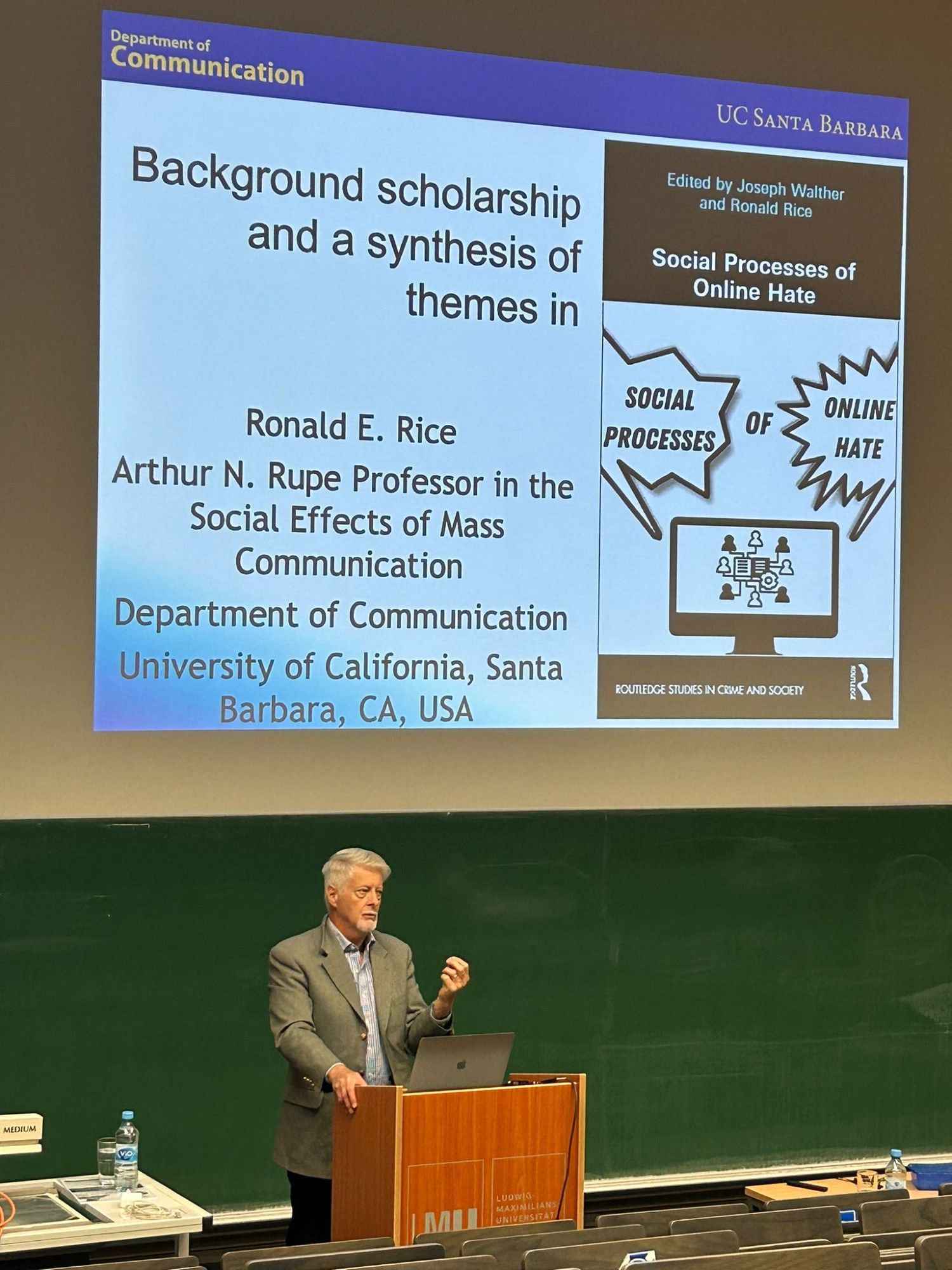 Prof. Ronald Rice steht in einem Hörsaal der LMU München. Hinter ihm ist eine Präsentation auf die Wand projiziert, die das Cover seines neuen Buches "Social Processes of Online Hate" zeigt.