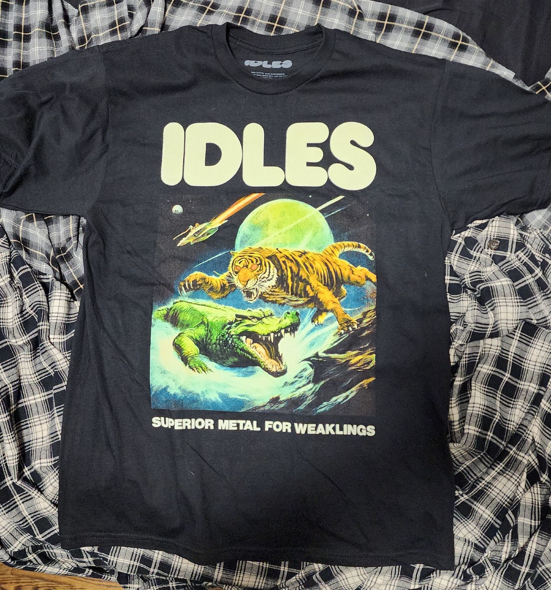 A photo of a black t-shirt that says IDLES on the top and SUPERIOR METAL FOR WEAKLINGS on the bottom. In the middle there's an image of a tiger jumping over an alligator while a flying saucer sails by in outer space in the background.