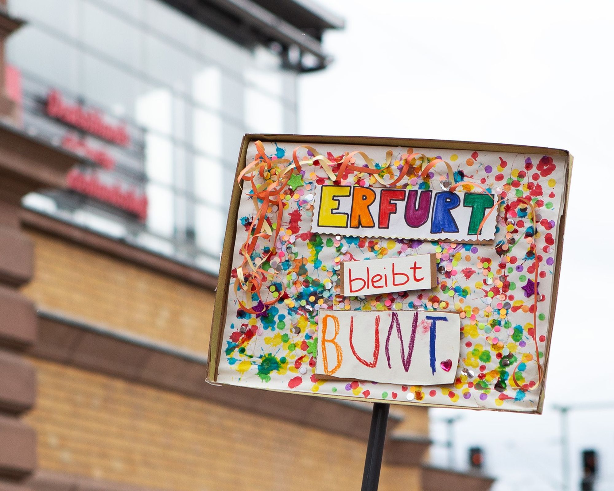 Demo gegen Rechts, in die Luft gehaltenes, bunt mit Luftschlangen und Konfetti verziertes Plakat, auf dem in allen Regenbogenfarben steht "Erfurt bleibt bunt."