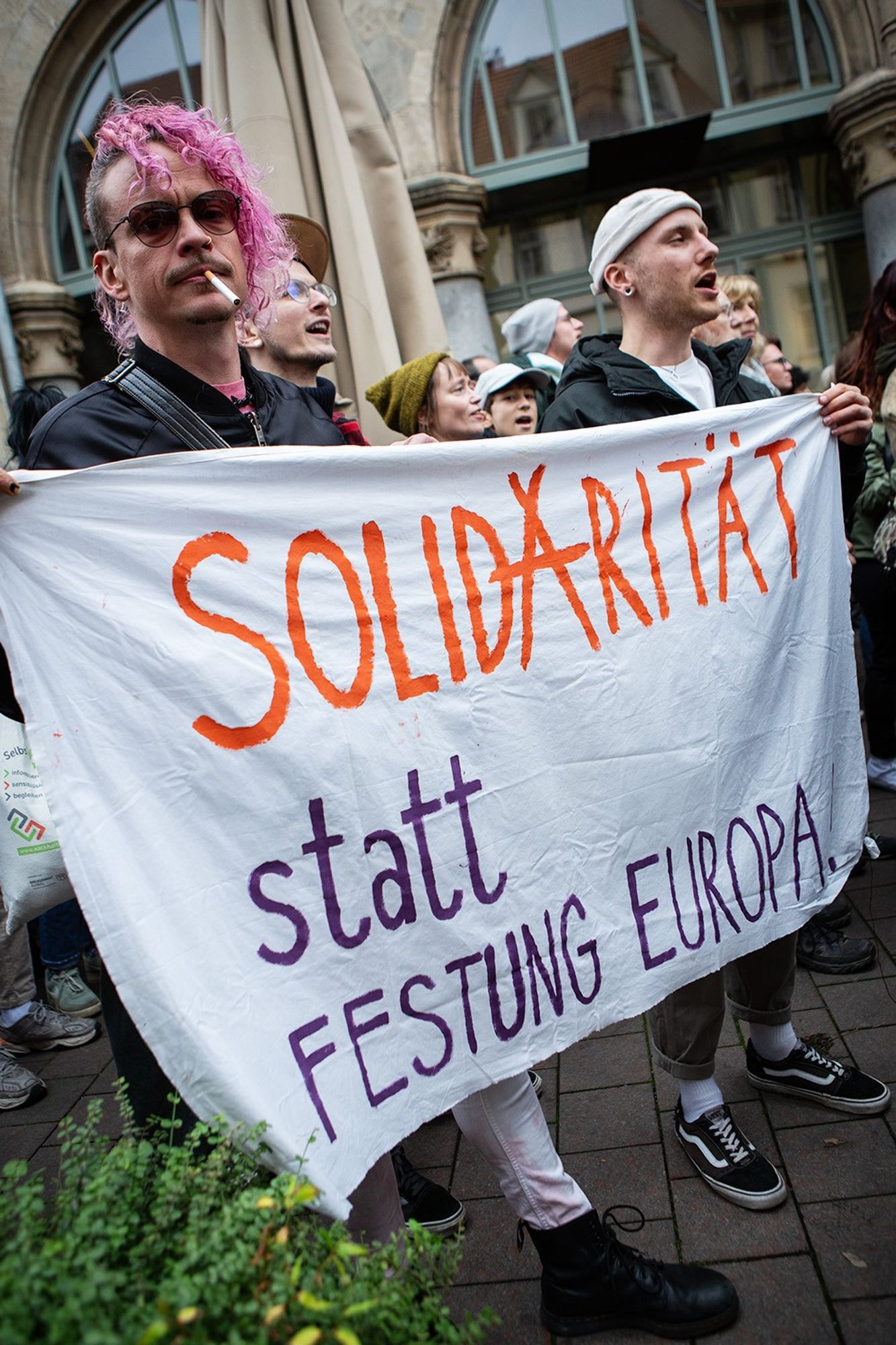 Demo gegen Rechts, im Demozug. Eine Gruppe überwiegend junger Leute rufen Parolen. Zwei Männer halten ein weißes Transparent "Solidarität statt Festung Europa". Der hintere in sportlicher Kleidung schaut nach vorn und ruft etwas. Der vordere ist modisch gekleidet, hat teils rasierte/teils pink gelockte Haare, trägt Sonnenbrille, Zigarette im Mundwinkel und schaut in die Kamera.