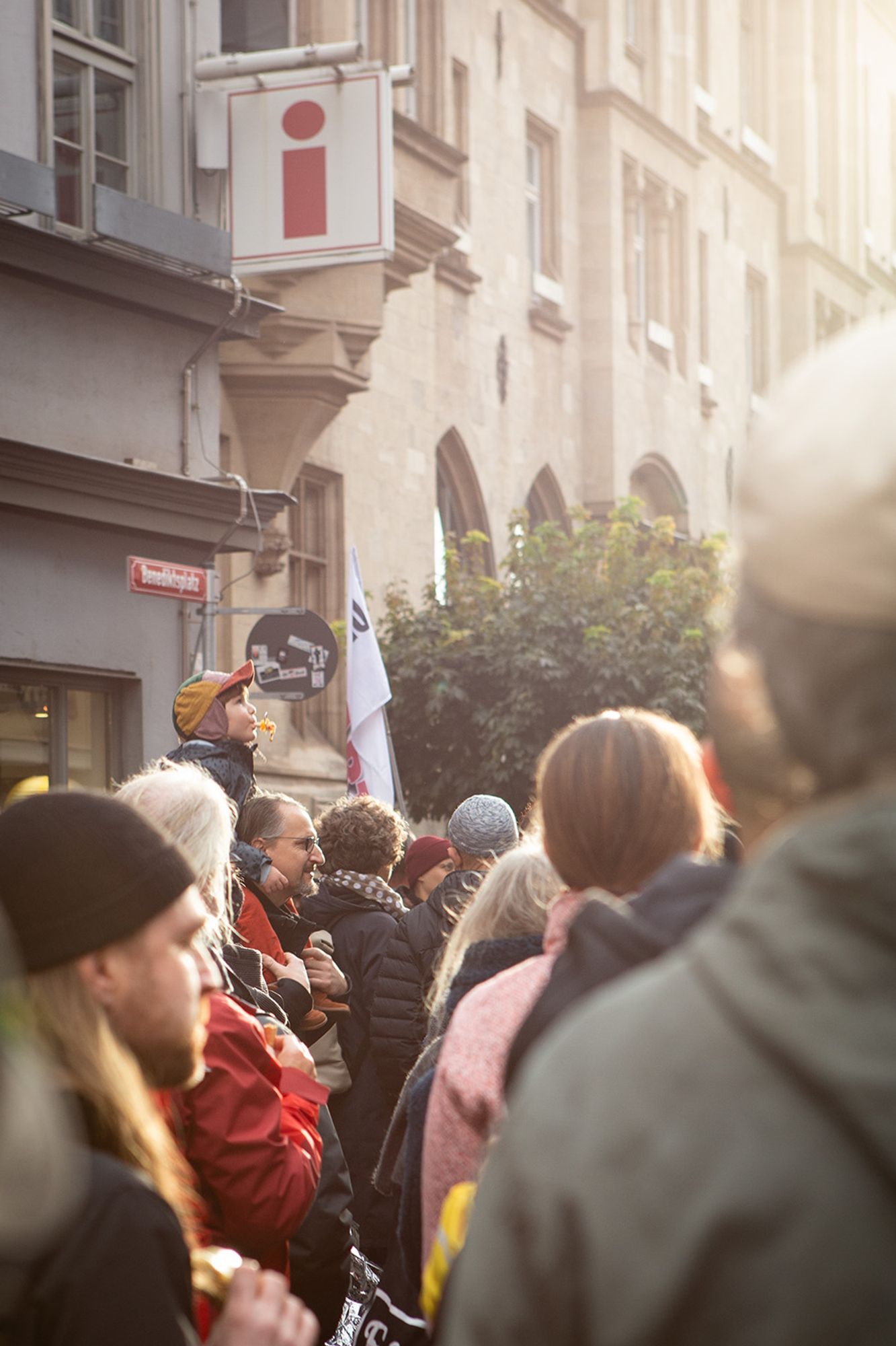 Demo gegen Rechts, im Demozug. Viele Menschen von hinten in einer schmalen Straße, Gegenlicht. Etwas links der Bildmitte ein etwas älterer Mann im Profil. Er trägt ein kleines Kind auf den Schultern. Das Kind trägt eine bunte Mütze und bläst vergnügt in eine gelbe Trillerpfeife.