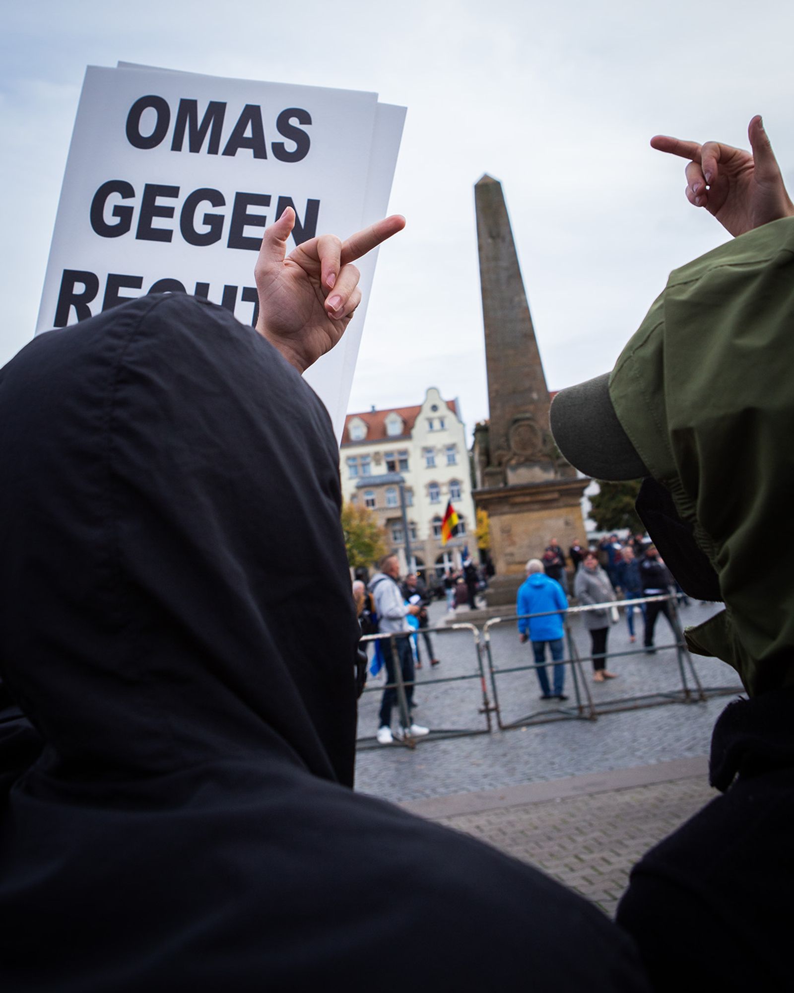 Demo gegen Rechts, im Demozug. Im Vordergrund ein Schild der "Omas gegen Rechts" und zwei vermummte Gestalten von hinten, die beide den Stinkefinger zeigen. Im Hintergrund eine Absperrung, dahinter ein Teil des Domplatzes Erfurt und vereinzelte Anhänger der AFD, die dort eine Demo abhalten.