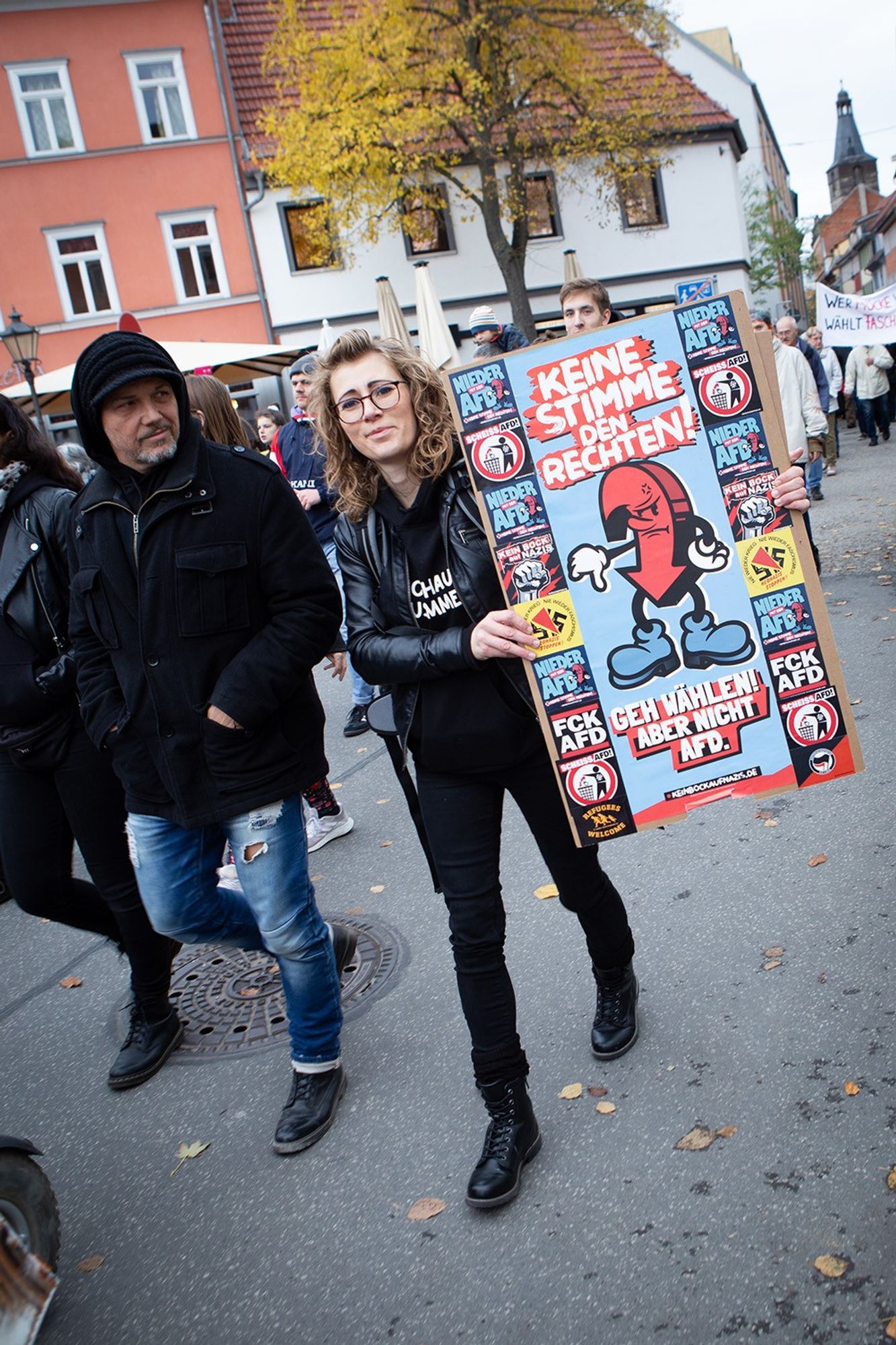 Demo gegen Rechts, im Demozug. Eine junge Frau, schwarz gekleidet mit schulterlangen, lockigen Haaren und Brille schaut in die Kamera und hält ein Schild auf dem steht "Keine Stimme den Rechten! Geh wählen! Aber nicht AFD." Neben ihr läuft ein ebenfalls schwarz gekleideter Mann, der zu ihr schaut.