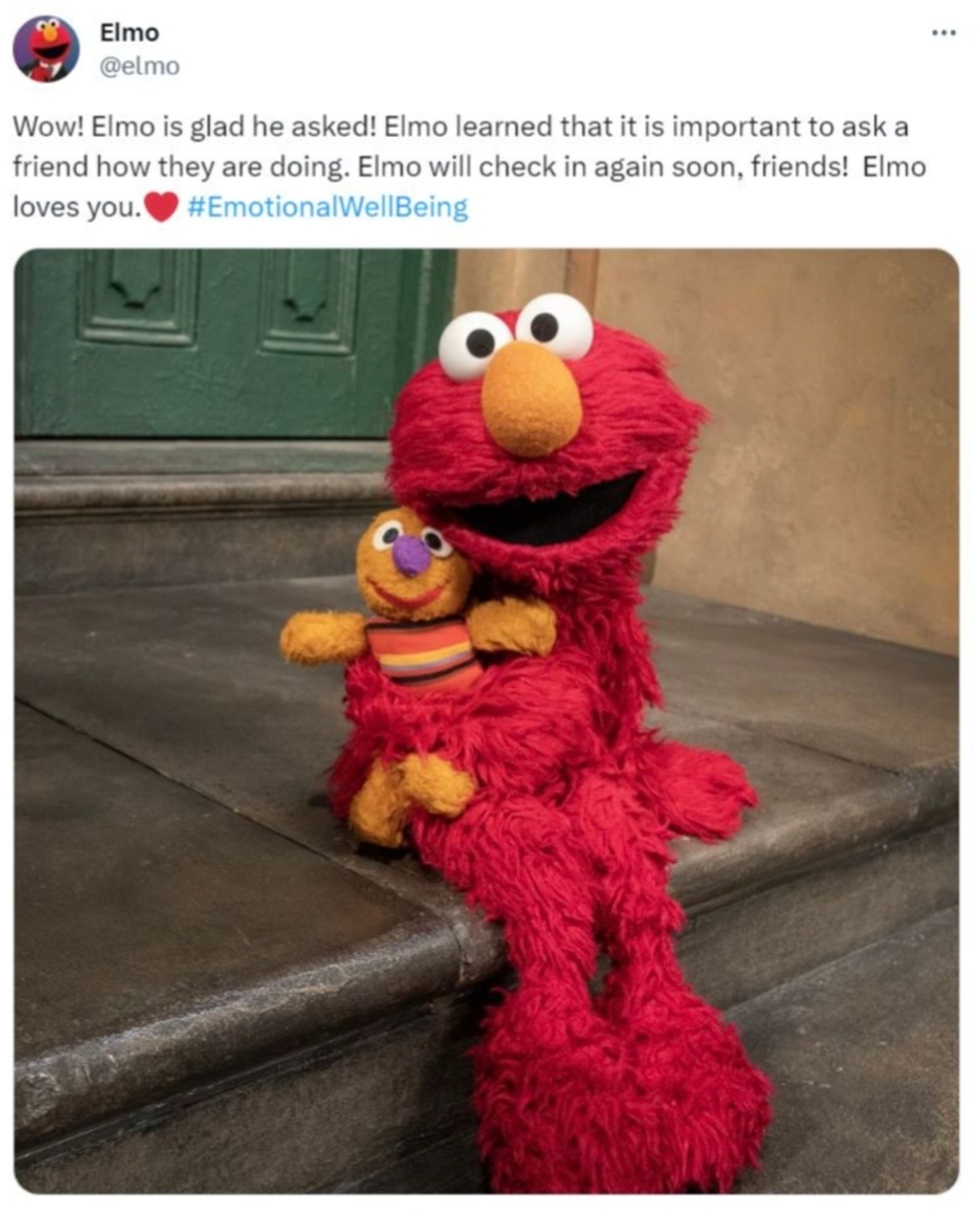 Elmo seated on a front stoop, holding a toy muppet, under an Elmo tweet that reads "Wow! Elmo is glad he asked! Elmo learned that it is important to ask a friend how they are doing. Elmo will check in again soon, friends! Elmo loves you. ❤️ #EmotionalWellBeing "