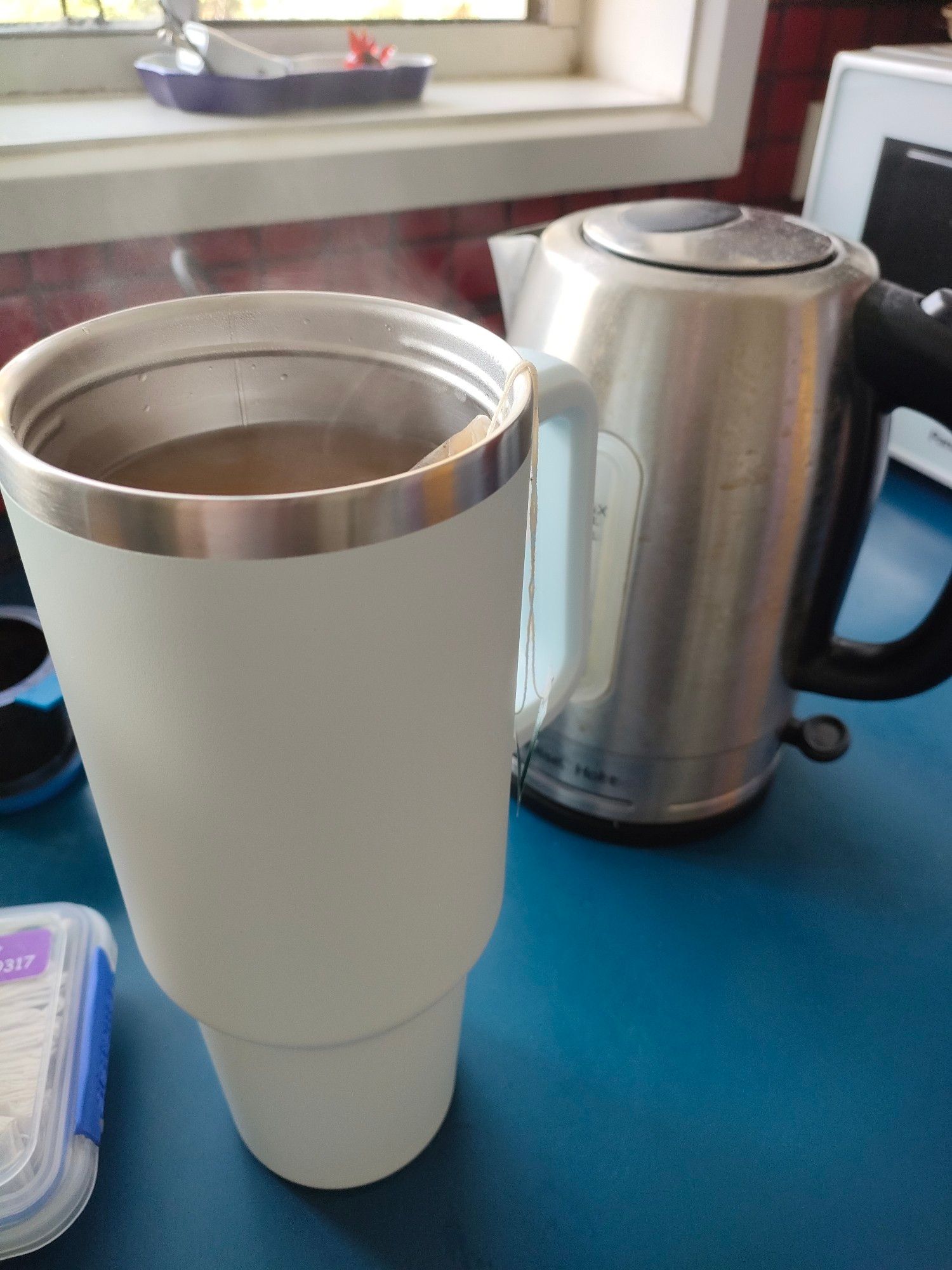 A 1.1 litre insulated travel mug full of hot tea sits on a kitchen bench next to a full-sized kettle. The kettle looks slightly smaller than the mug (it does hold more than the mug does though).