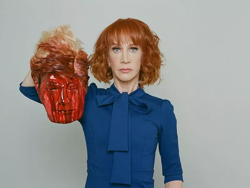Kathy Griffin- a pale white woman with red hair and wearing a blue dress- holding up in her left head a mask made to look like donald trump by the hair as if he has just been decapitated. Melania claimed it confused Barron who thought his dad really died which shows that kid is stupid af too