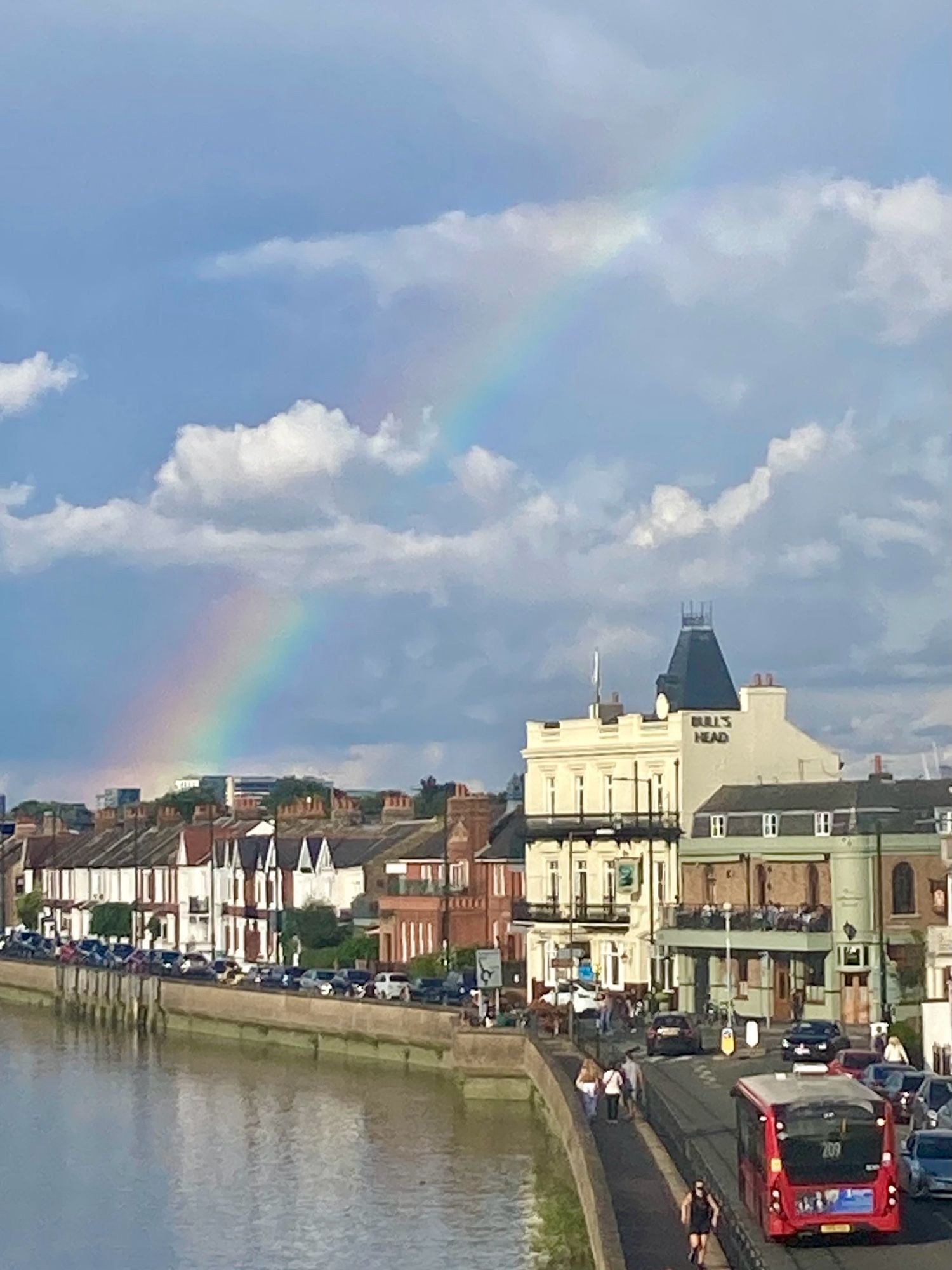 A rainbow in blue and cloudy sky above the Batnes riverfront brightly lit by afternoon sun, a bus drives towards the junction with Barnes High Street