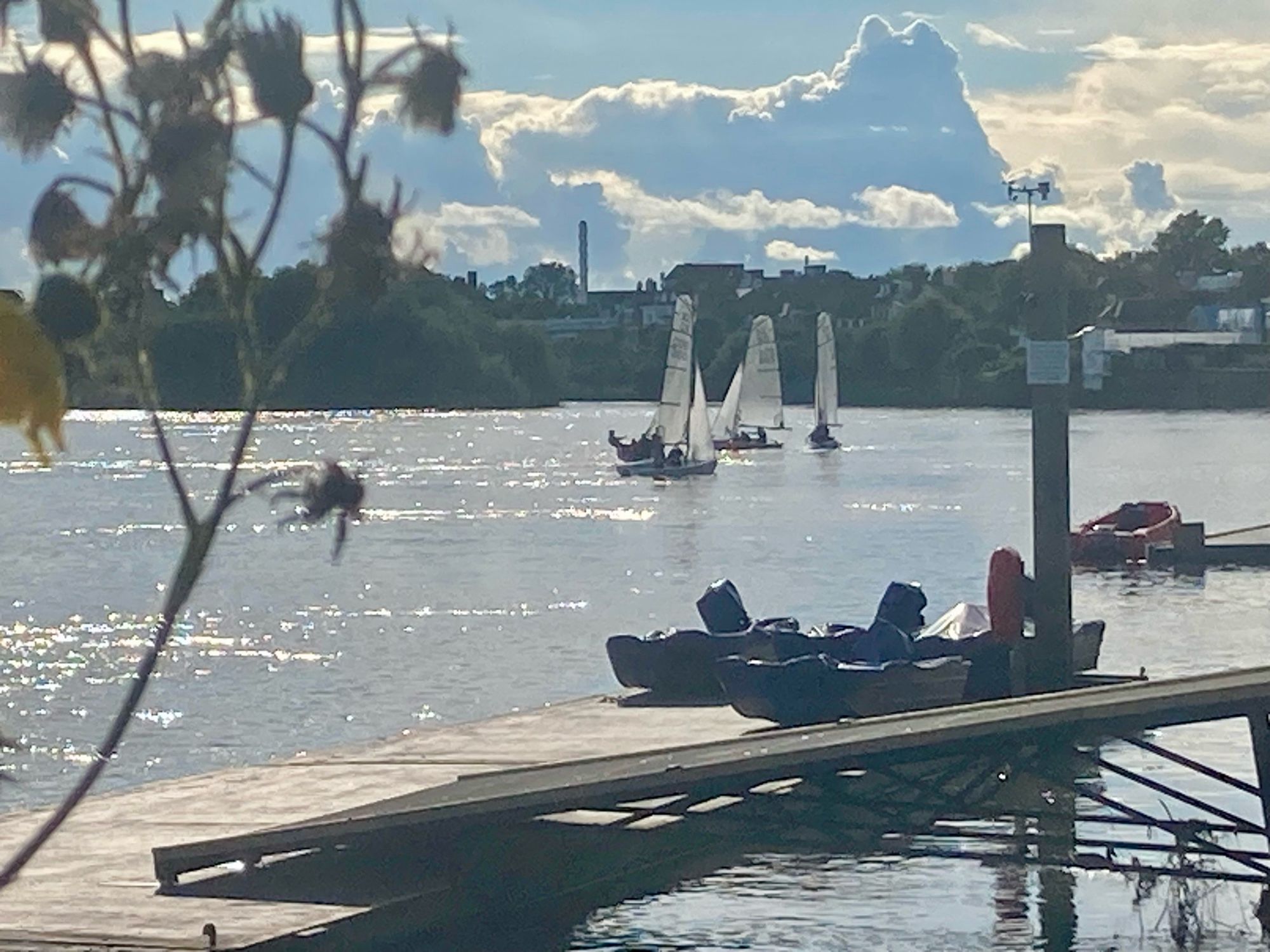 Sunlight dazzling on the water, the jetty and some sailing dinghies, trees on distant bank and towering clouds above the horizon