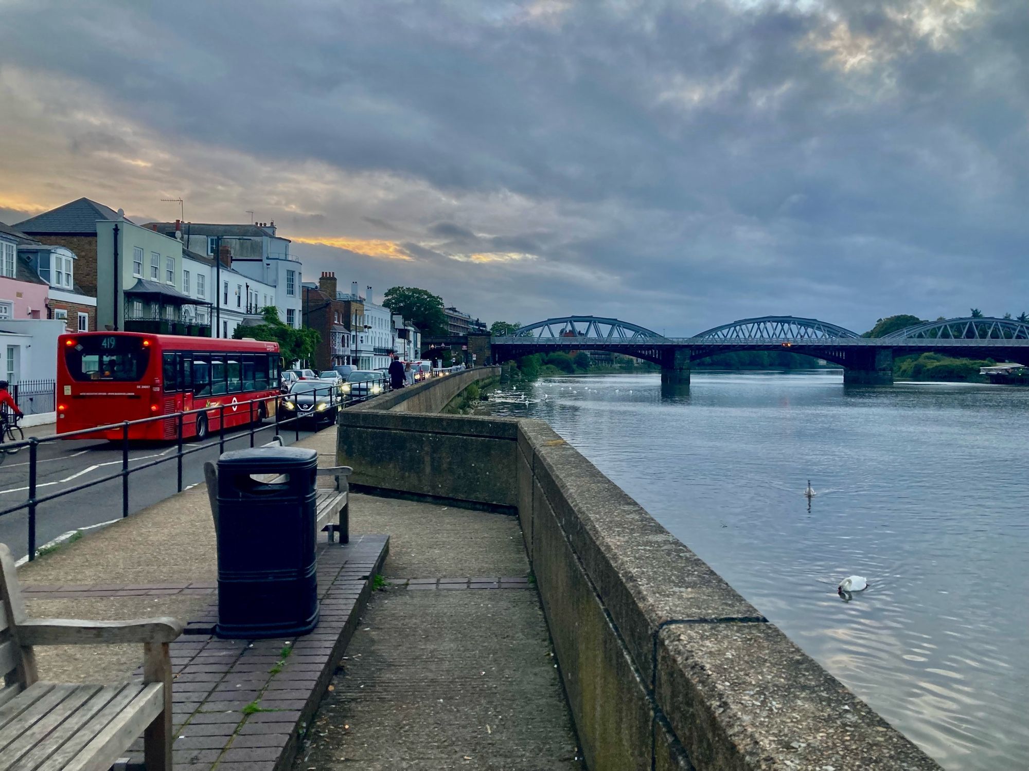 A number 419 single-decker red London bus drives next to the Thames with a backdrop of house fronts and zigzagging parapet in foreground each receding in perspective towards Barnes Bridge