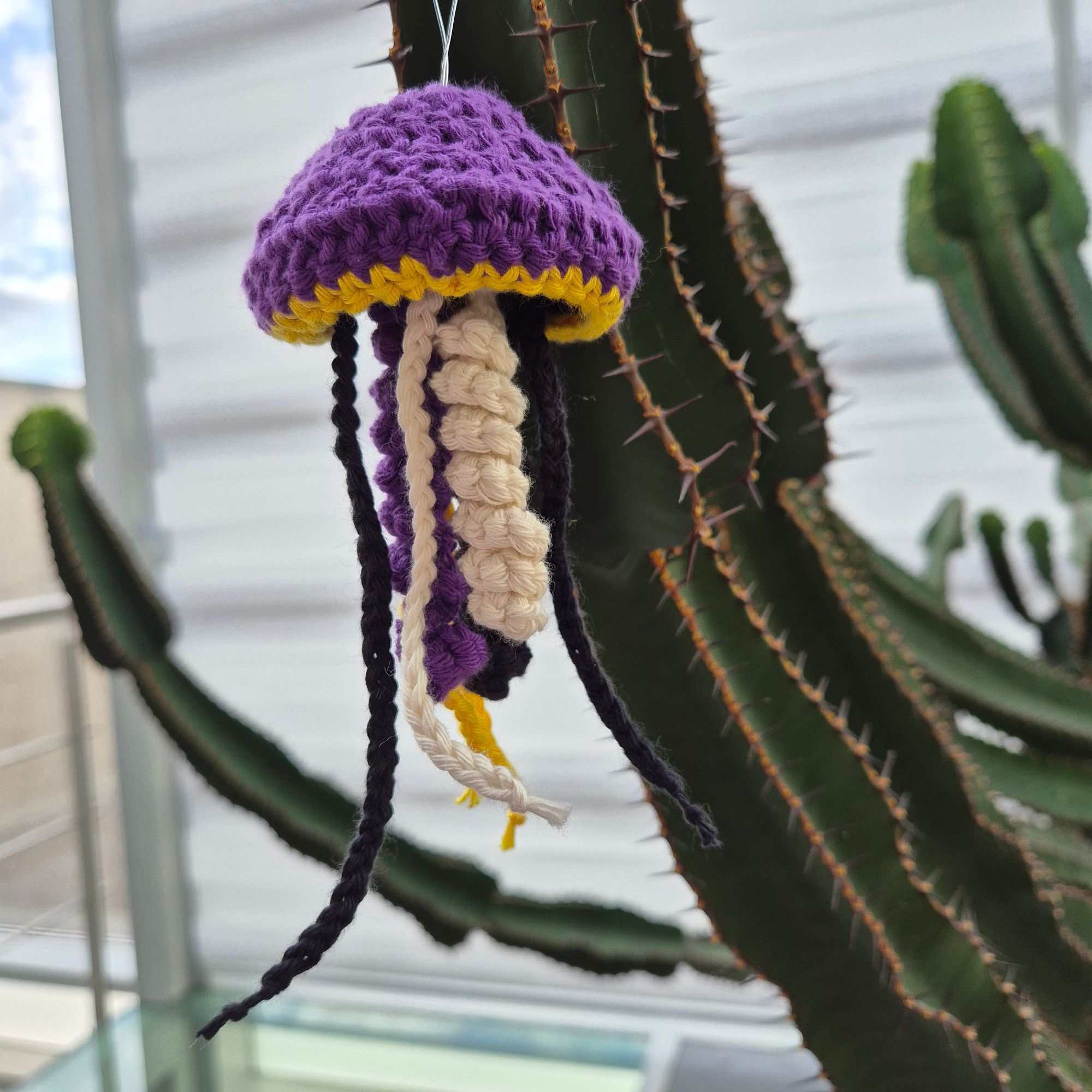 A crocheted cactus in purple, yellow, black, and white cotton is hanging from the arm of a large green cactus.