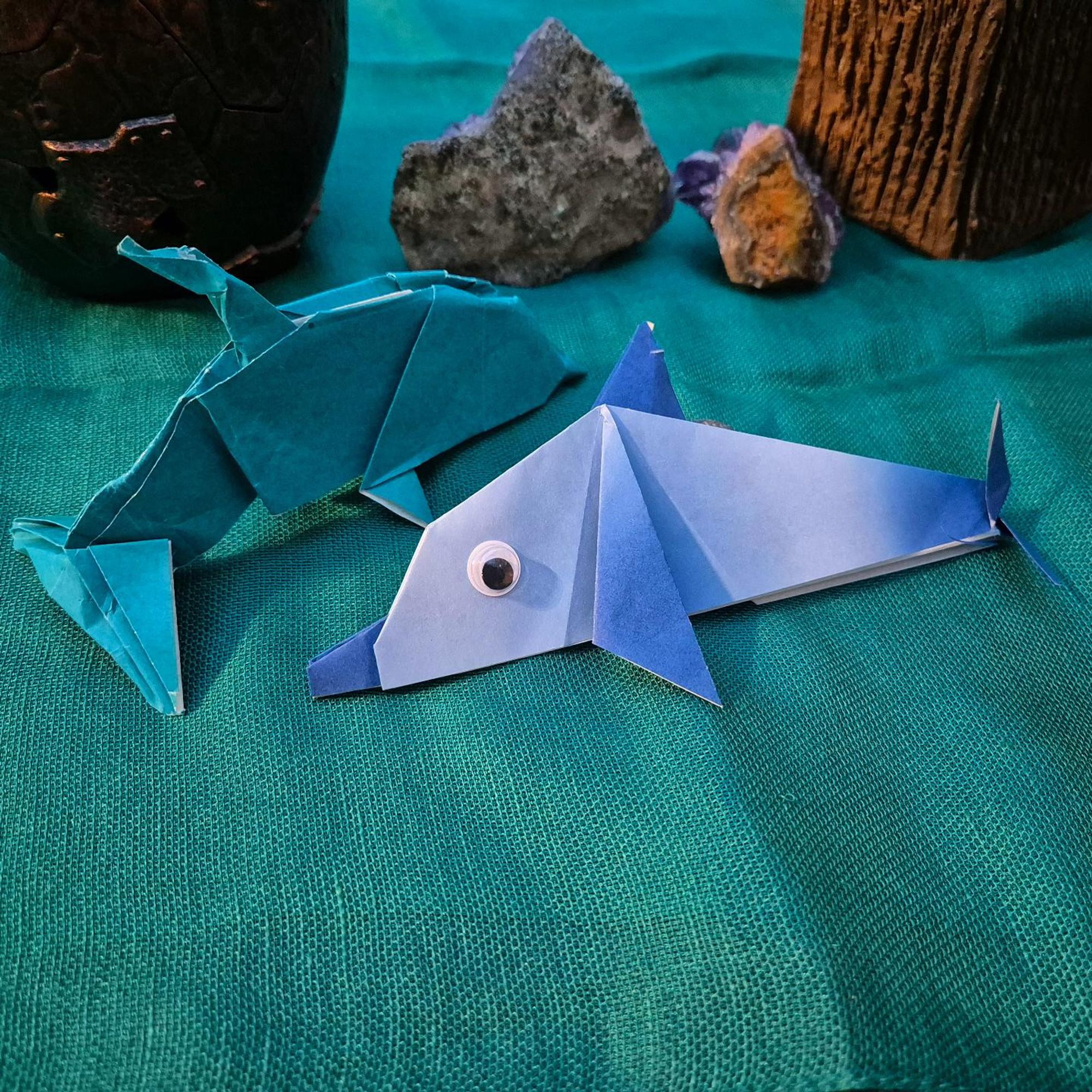 Photo of two different origami dolphins in different shades of blue, one of them looking into the camera with a googly eye. They are lying on a green fabric underground with the impression of rocks behind them.