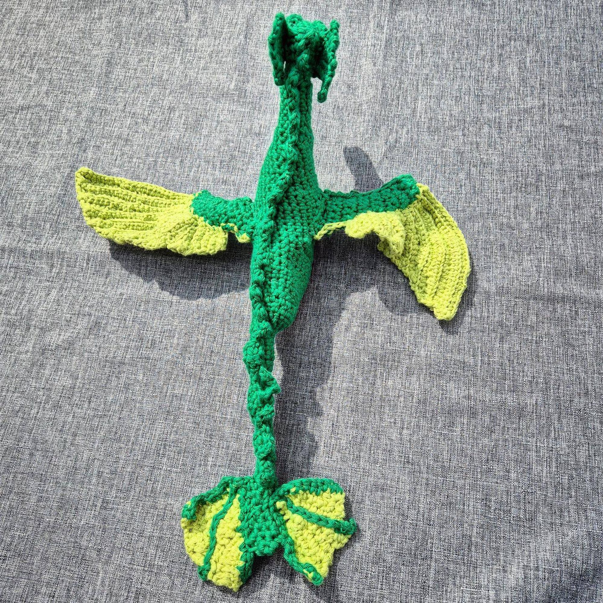 Photo of a crocheted sea dragon with a dark green body and light green details on its wings and tail, seen from the back.