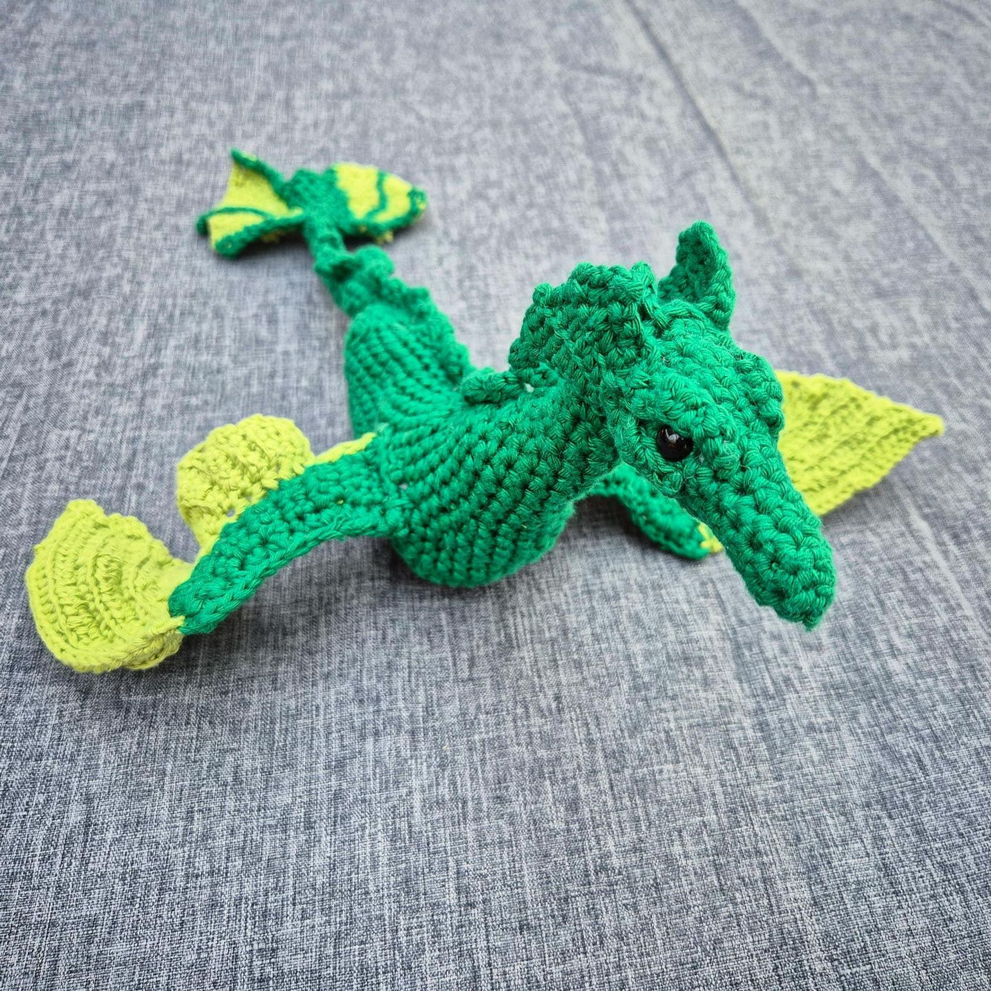 Photo of a crocheted sea dragon with a dark green body and light green details on its wings and tail, angled so one of its eyes looks into the camera.