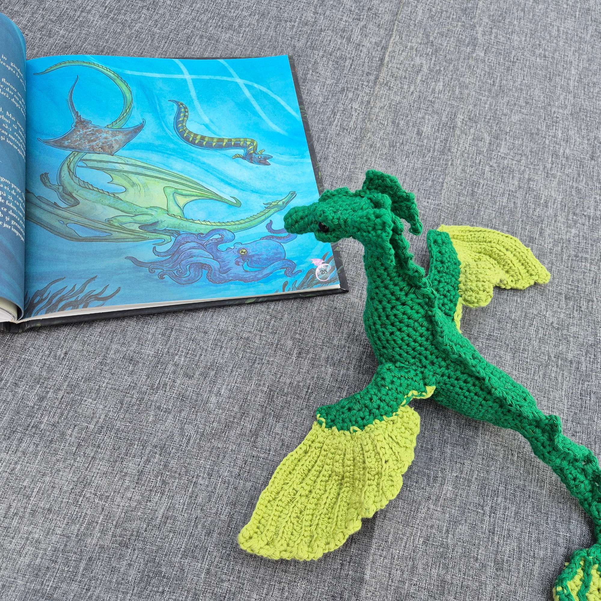 Photo of a crocheted green dragon in front of a colourful page of a picture book depicting a similar dragon accompanied by a ray, a sea snake, and an octopus.