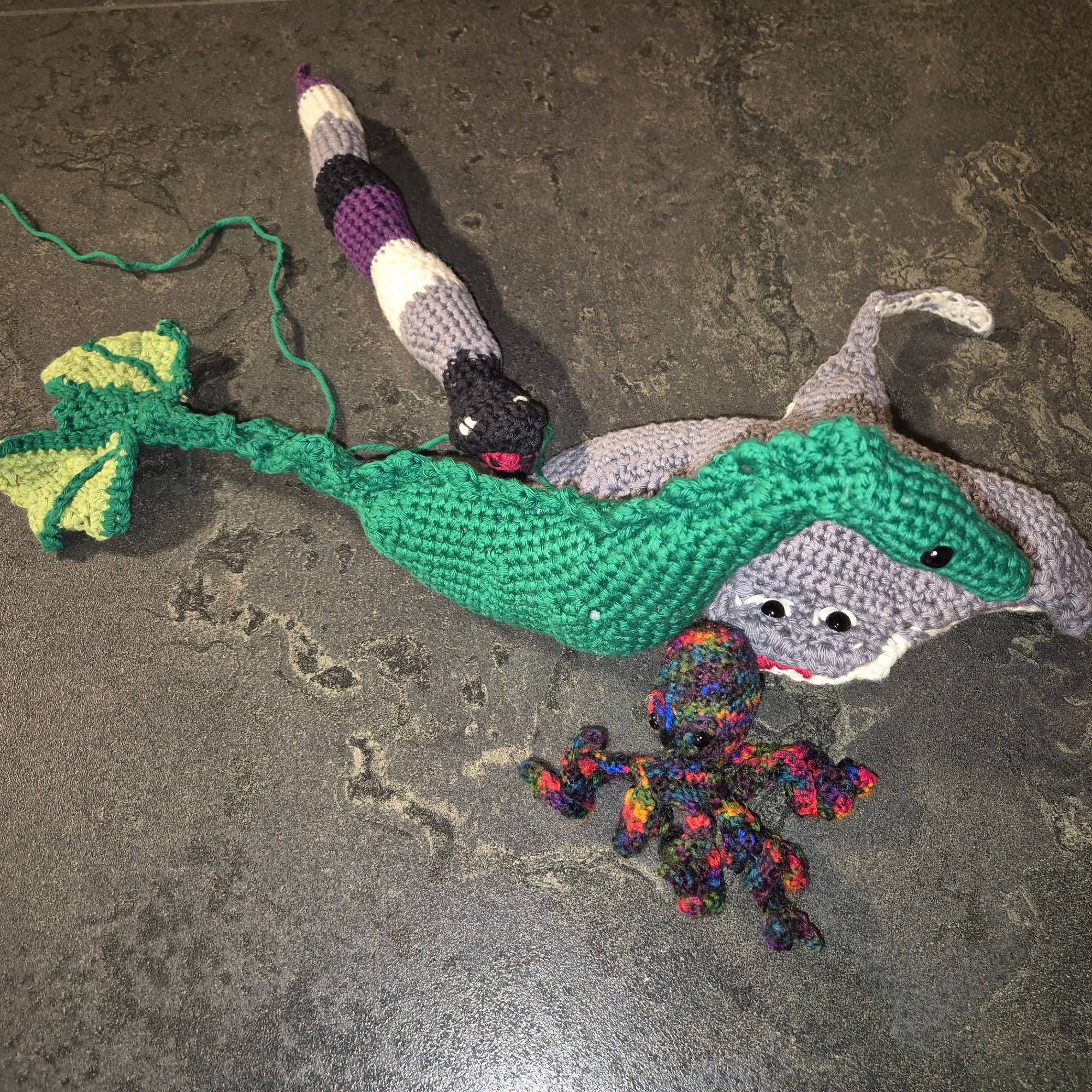 Photo of a group of crocheted animals: a green dragon without legs or wings but with a mermaid tail, supported by a grey manta ray, a rainbow octopus, and a sea snake in the colours of the asexual flag, on a grey background.