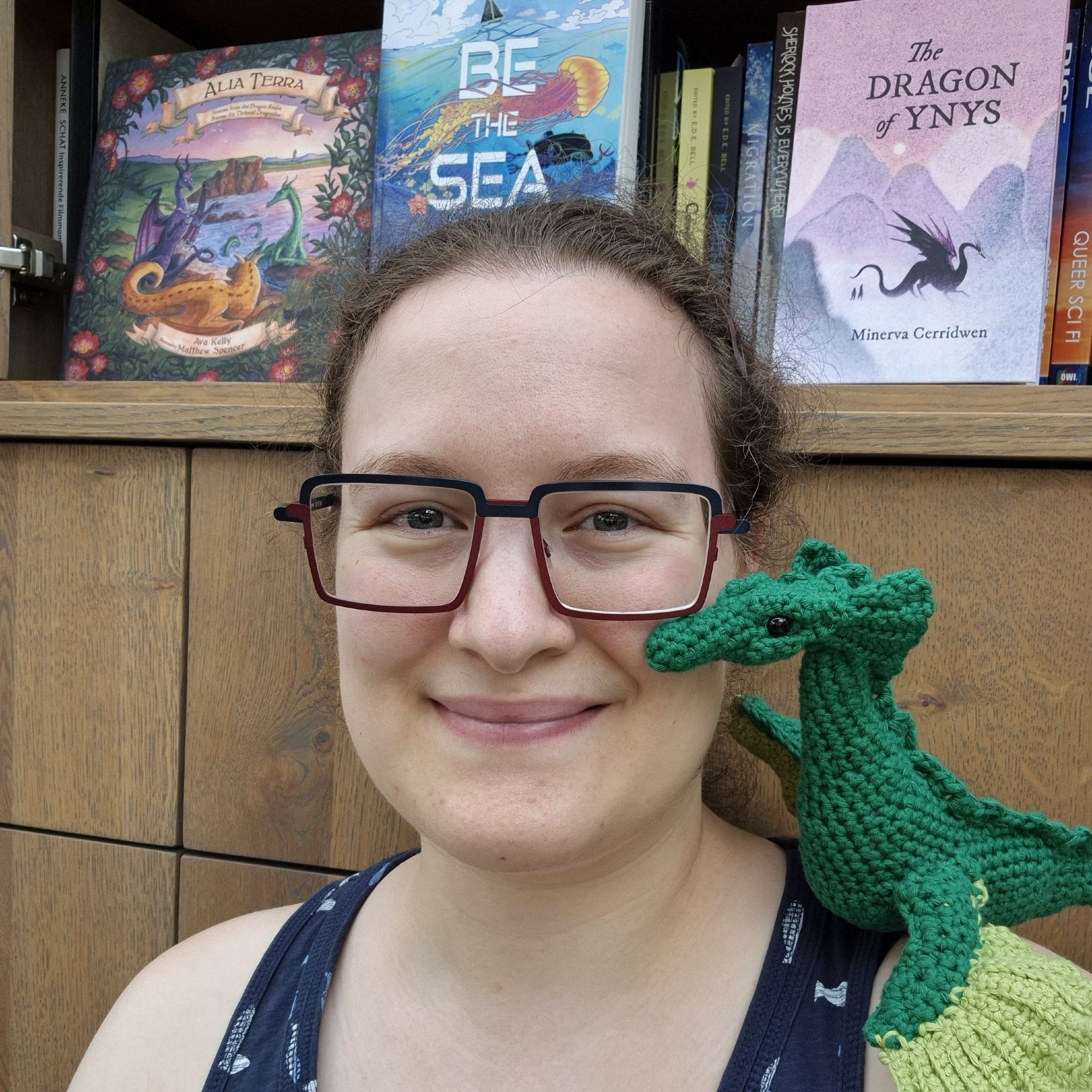 Photo of a smiling person with glasses wearing a navy tank top with fish print, who has a green crocheted dragon sitting on xyr left shoulder. In the background 3 books are on display: Alia Terra, Be the Sea, and The Dragon of Ynys.