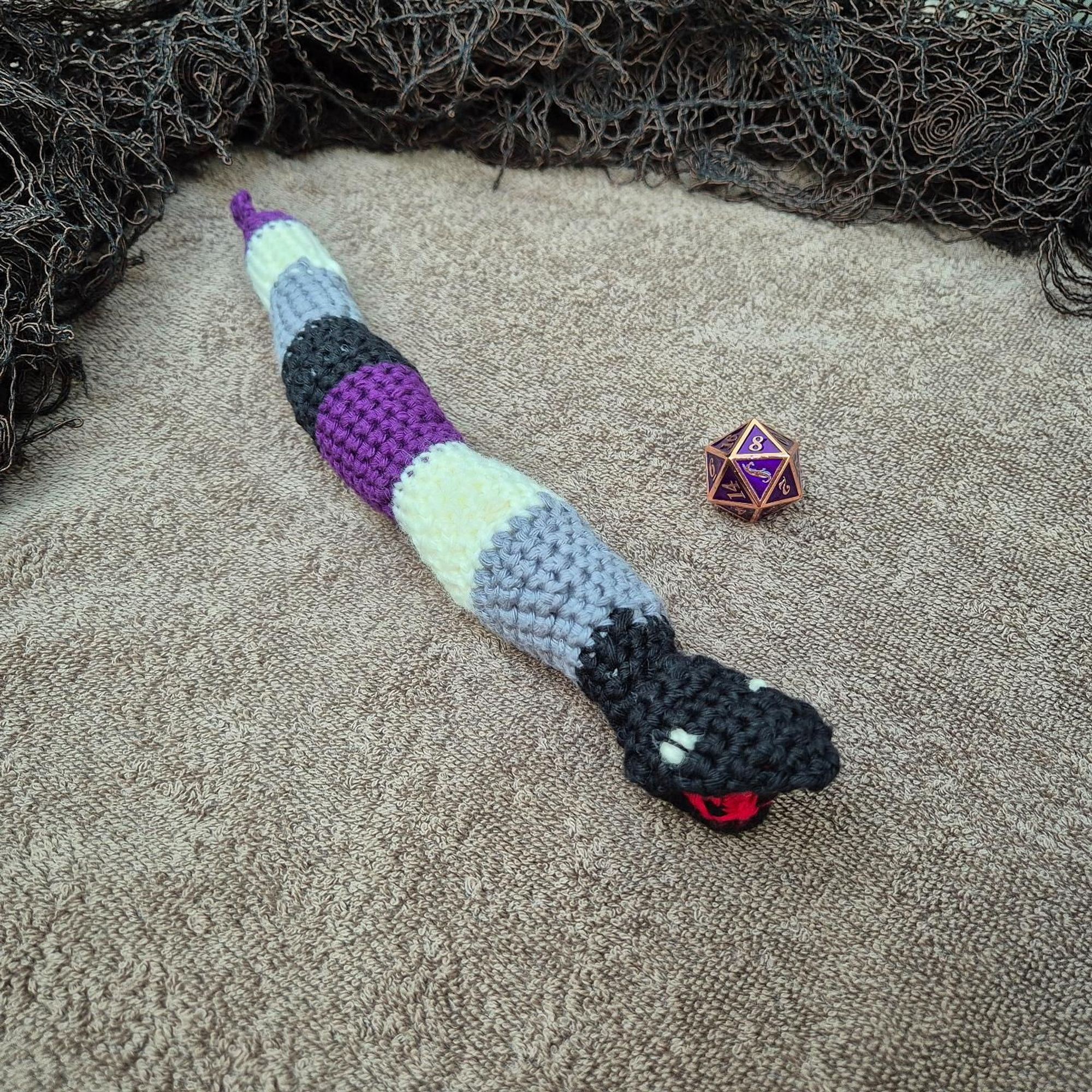 Photo of a crocheted sea snake in black, grey, white and purple, next to a purple d20 die, on a brown towel that reminds of a sandy beach, with dark fabric around it that looks like dried algae.