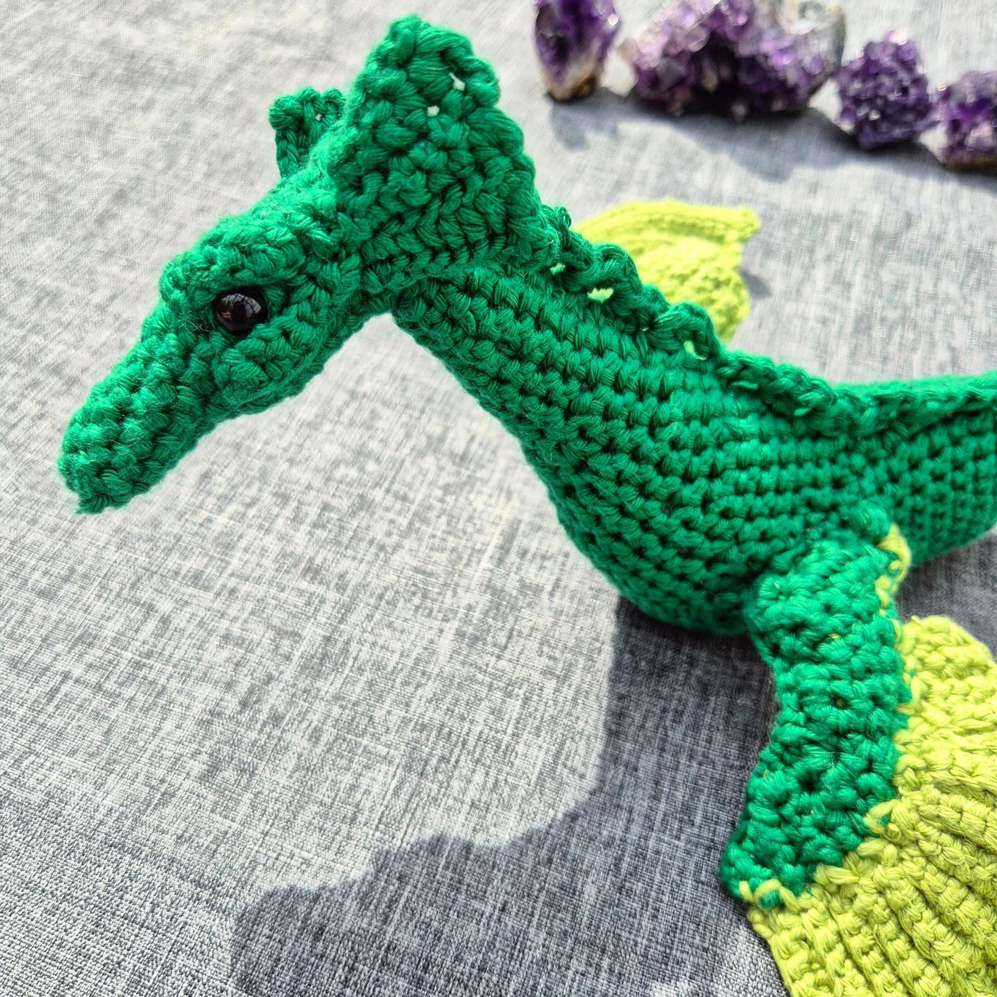 Close-up photo of the head of a green crocheted sea dragon, with amethyst crystals in the background.