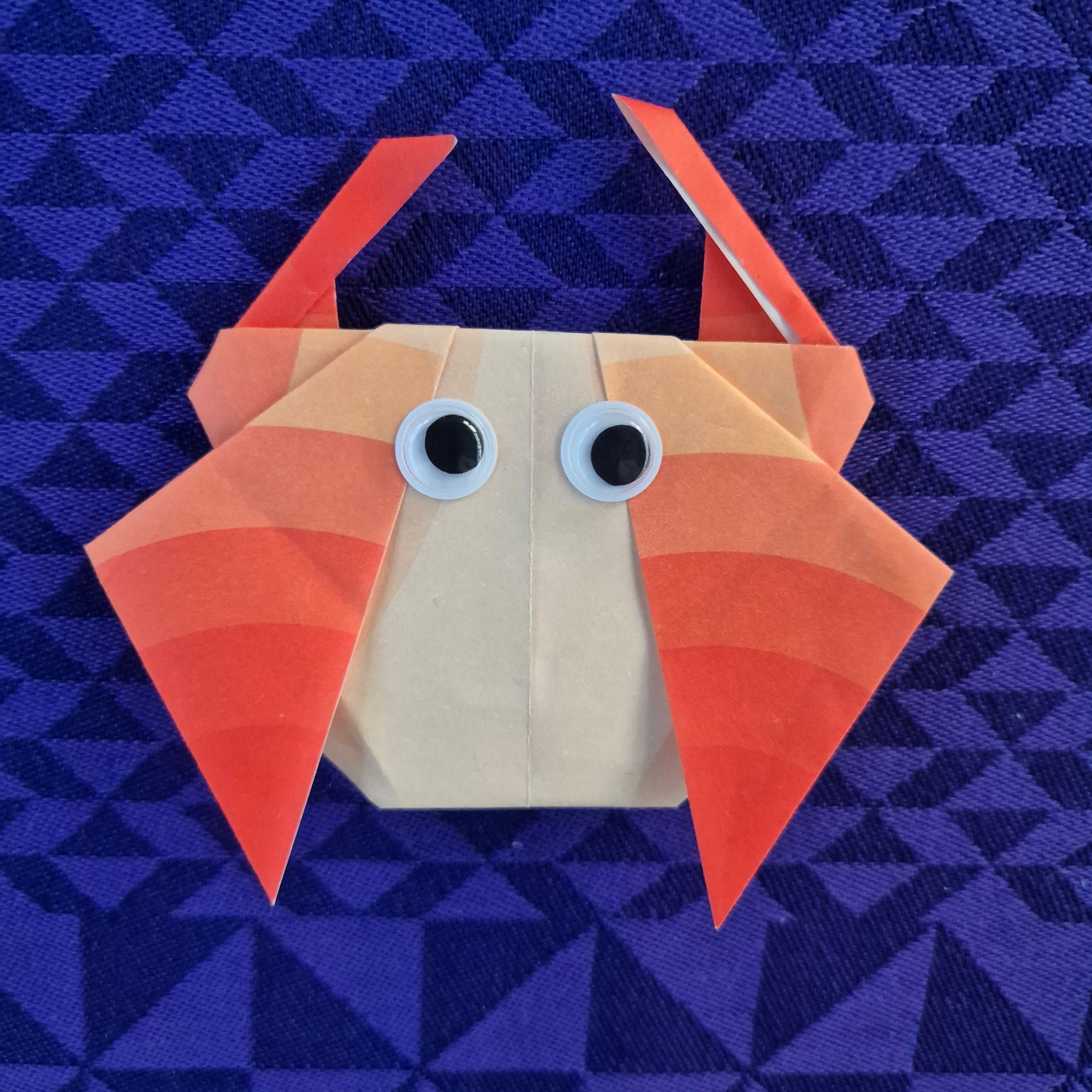 Photo of an origami crab made of paper patterned in different shades of orange, with googly eyes stuck to the top of the crab. It lies on top of a dark purple patterned tablecloth.