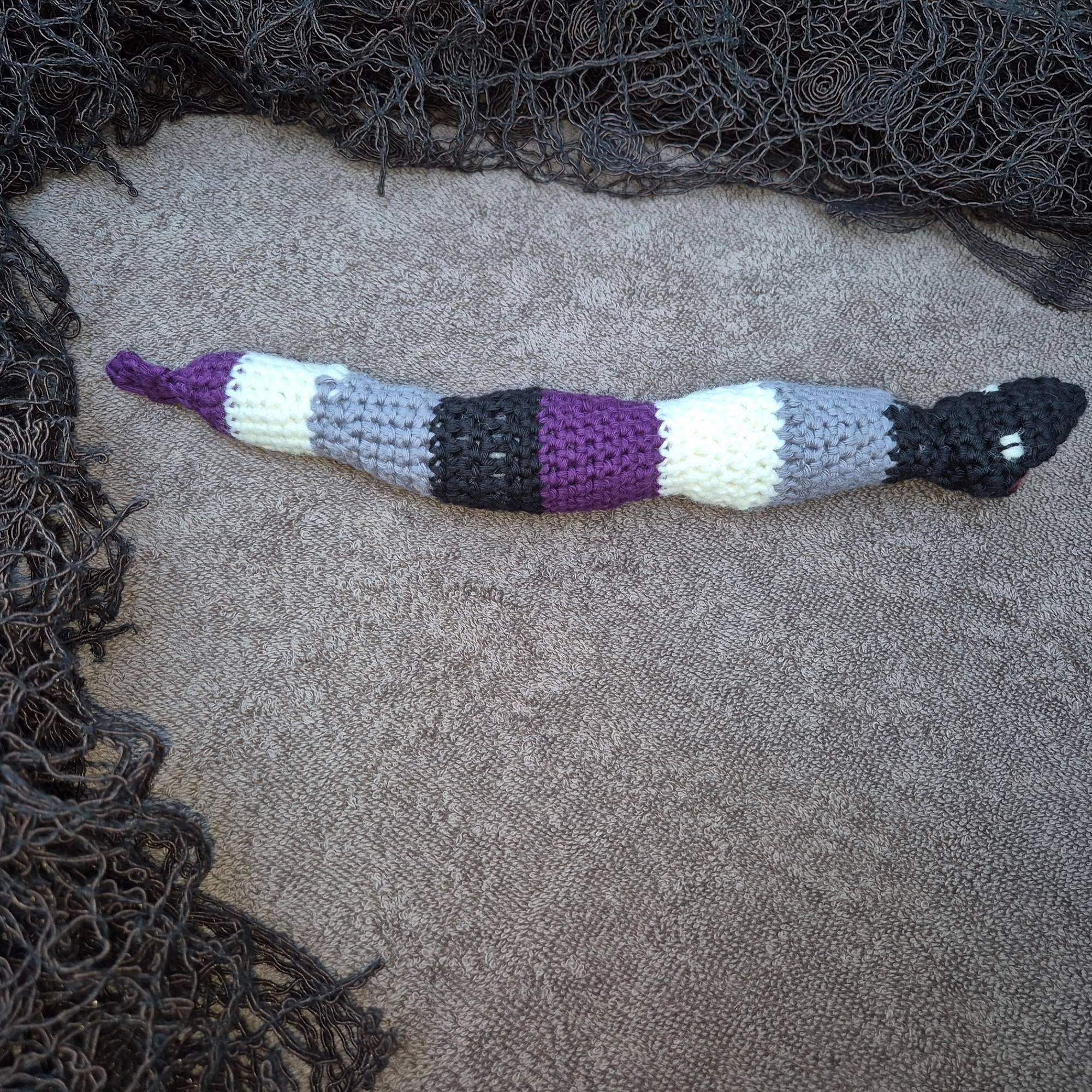 Photo of a crocheted sea snake in black, grey, white and purple, on a brown towel that reminds of a sandy beach, with dark fabric around it that looks like dried algae.