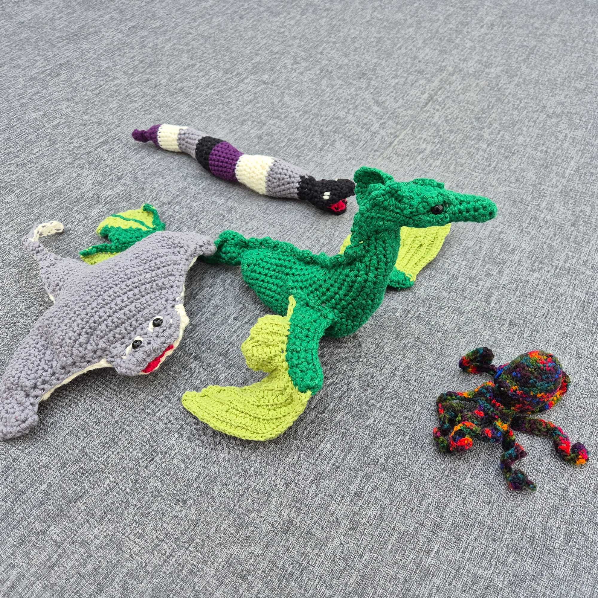 Photo of a crocheted grey ray, green dragon, sea snake in the colours of the asexual flag, and rainbow octopus, set up in the position as seen in the picture book on the previous photo.