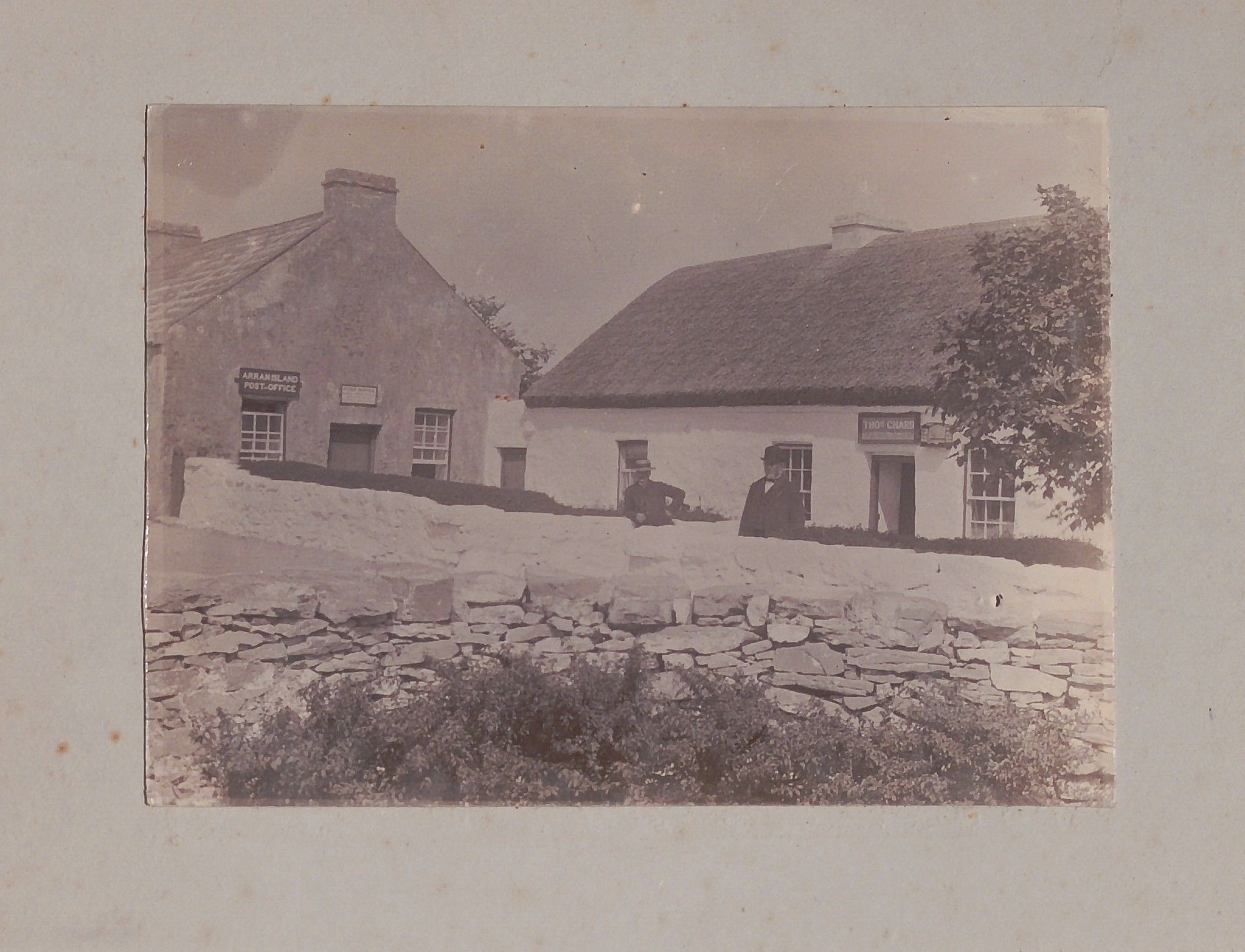 One image depicts the 'Arran Island Post Office' and was taken in Cill Rónáin, Árainn. The building directly to the right was known as Lios Aengus and today is a Bed & Breakfast, the Dormer House. The right half of the frame is occupied by a shop owned by Thomas Chard, a Protestant born c. 1827 in Co. Cork, who, by 1911 was still living in Cill Rónáin. He first came from Inishbofin to Aran in the late 1850s to take up a post as a schoolteacher. The age and dress of the man in the photograph suggests it could be a portrait of Thomas Chard himself outside his establishment.