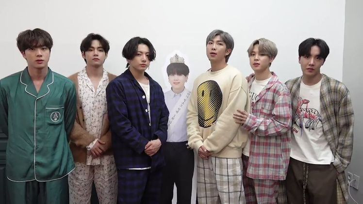 The image shows six BTS members standing in a casual setting, dressed in pajamas and relaxed clothing. In the center of the group is a standee of Yoongi (Suga), dressed in a white suit with a sash and crown, giving him a celebratory, regal appearance. The other members are gathered around the standee, each expressing a neutral look, creating a relaxed atmosphere.