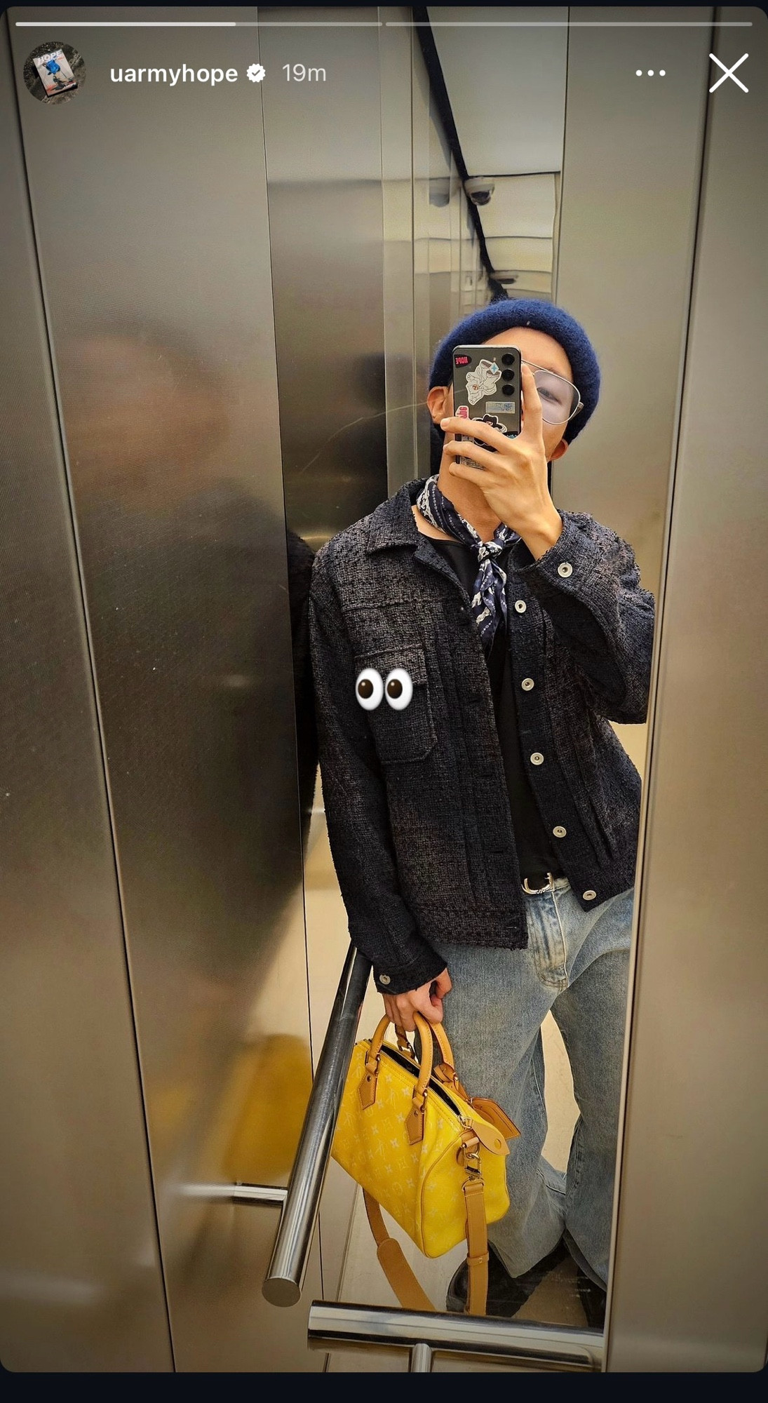 Hobi taking a mirror selfie, wearing a textured dark jacket, loose light-blue jeans, and a navy-blue beret. He is holding a bright yellow handbag and has a phone case decorated with stickers. The selfie is taken in an elevator, and the reflection captures multiple angles of Hobi and the setting.