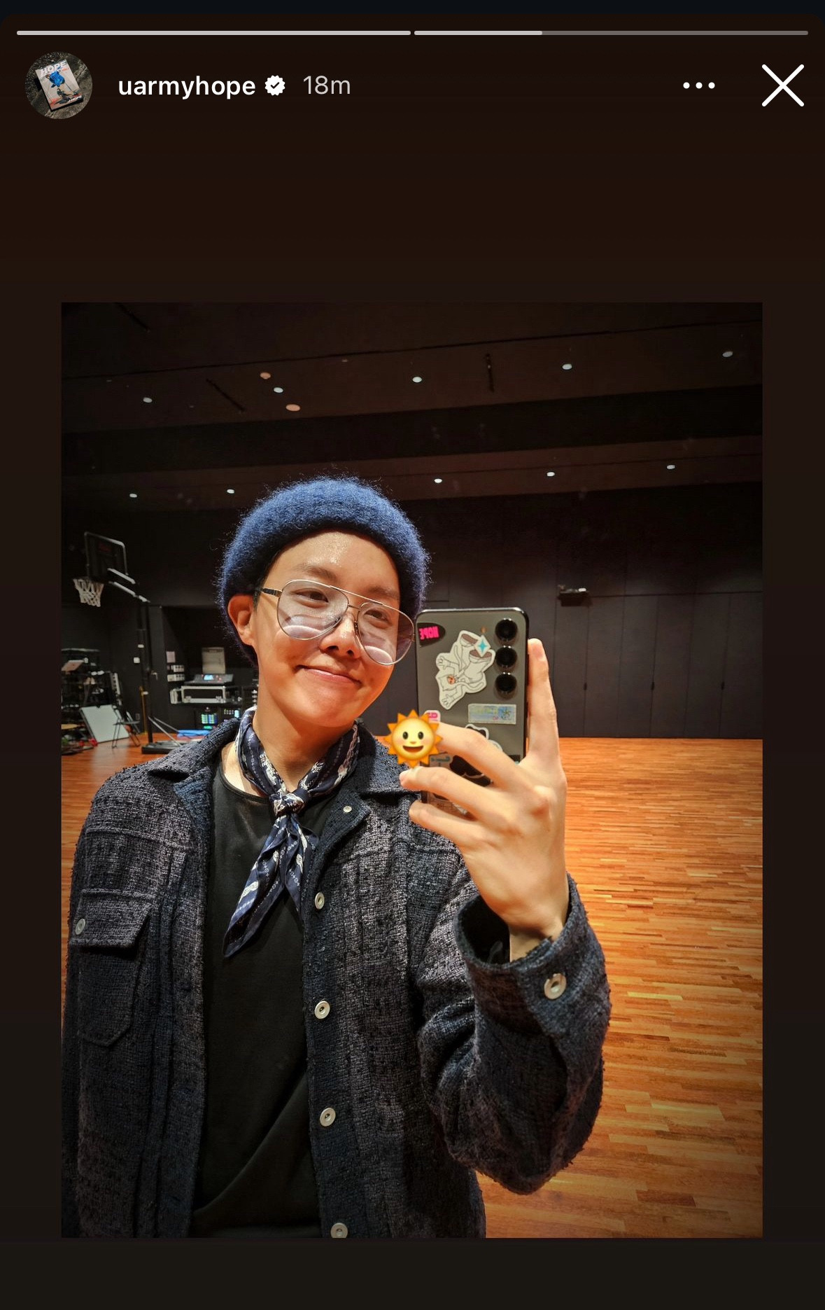 Hobi taking a mirror selfie, wearing a textured dark jacket, loose light-blue jeans, and a navy-blue beret. He is holding a phone decorated with stickers. 