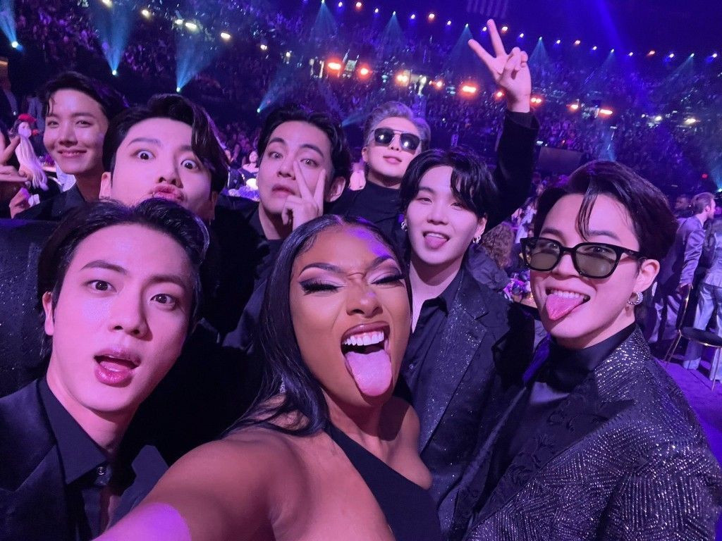 Meghan Thee Stallion and BTS making silly faces, all dressed in black, huddled together for a group selfie. 