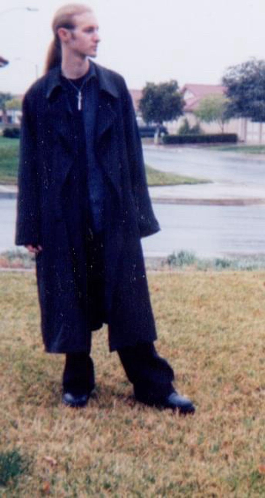 Photo of Draco, standing on a lawn of brown grass in a suburban neighborhood in Southern California. He has long dark blonde hair pulled back to a pony tail that falls down his back. He is looking off into the distance to give an air of mysterious aloofness. He’s wearing black boots, black pants, and a black t-shirt with a dark drey button up over it. A long black London Fog coat hangs off of his shoulders. A silver sword necklace from Hot Topic stands out from the black background of his shirt. He’s probably thinking about the next Type O Negative show he’s going to attend or maybe about the homework he needs to finish for a community college course. He’s definitely not embarrassed about having to ask his mom to take these photos for him.