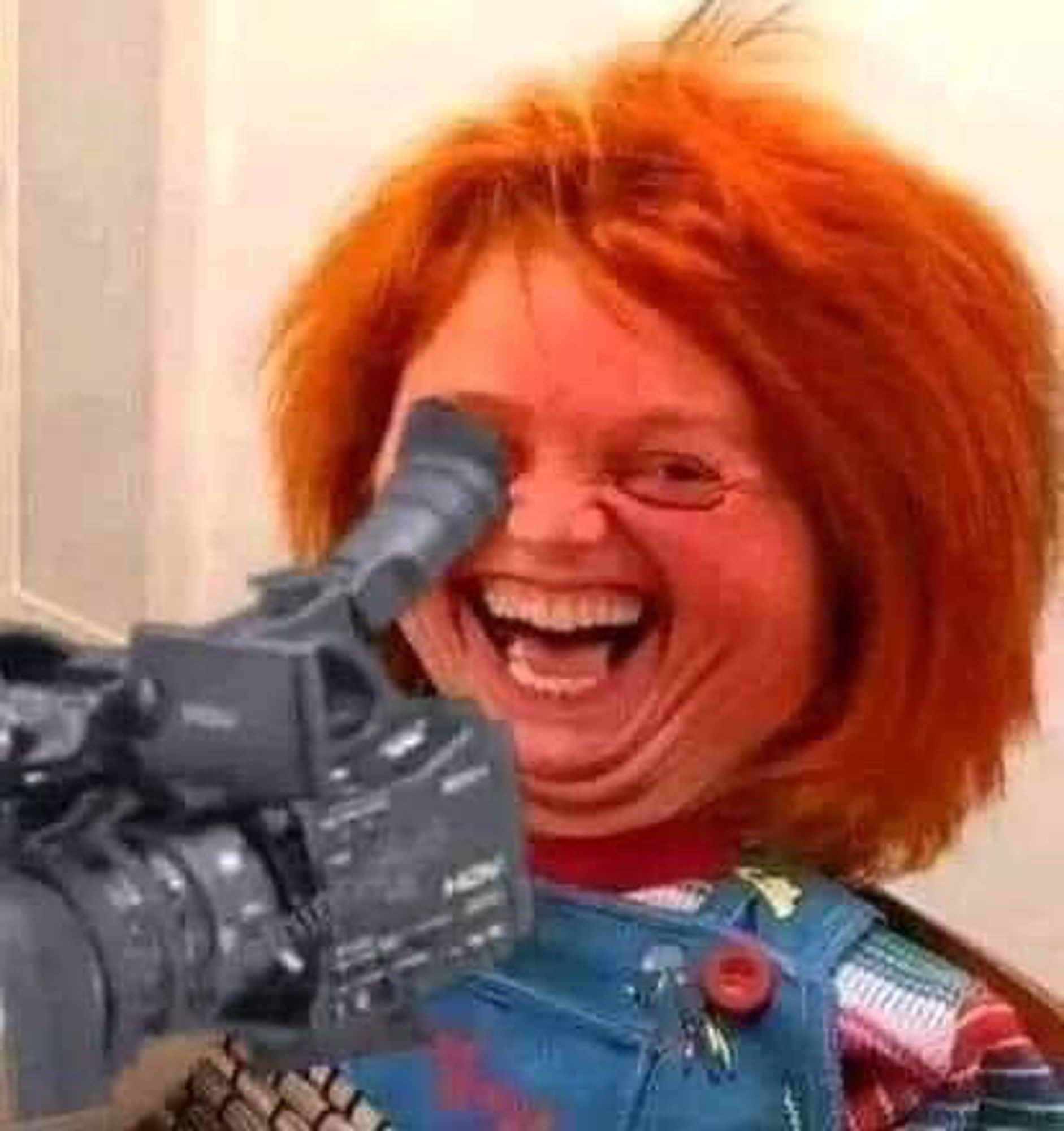 Chucky the Doll has been edited to have a huge ole human smile, he’s laughing. He’s cheesing. he is pointing a handheld camera just off frame to the left