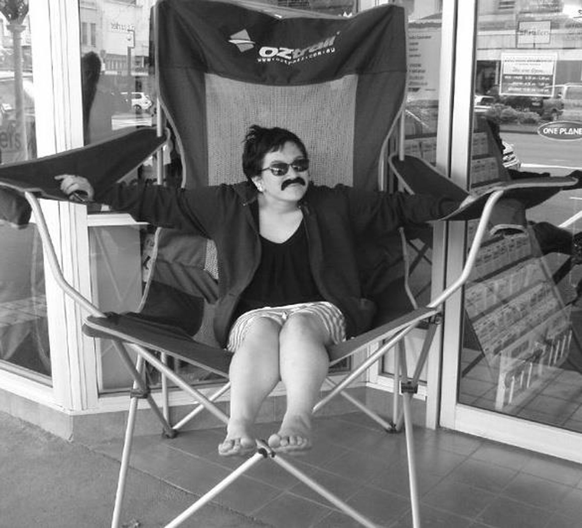 a black and white image of younger Puck in a giant promotional camping chair. They have a false moustache on and dark sunglasses. They are absolutely swallowed by the chair.